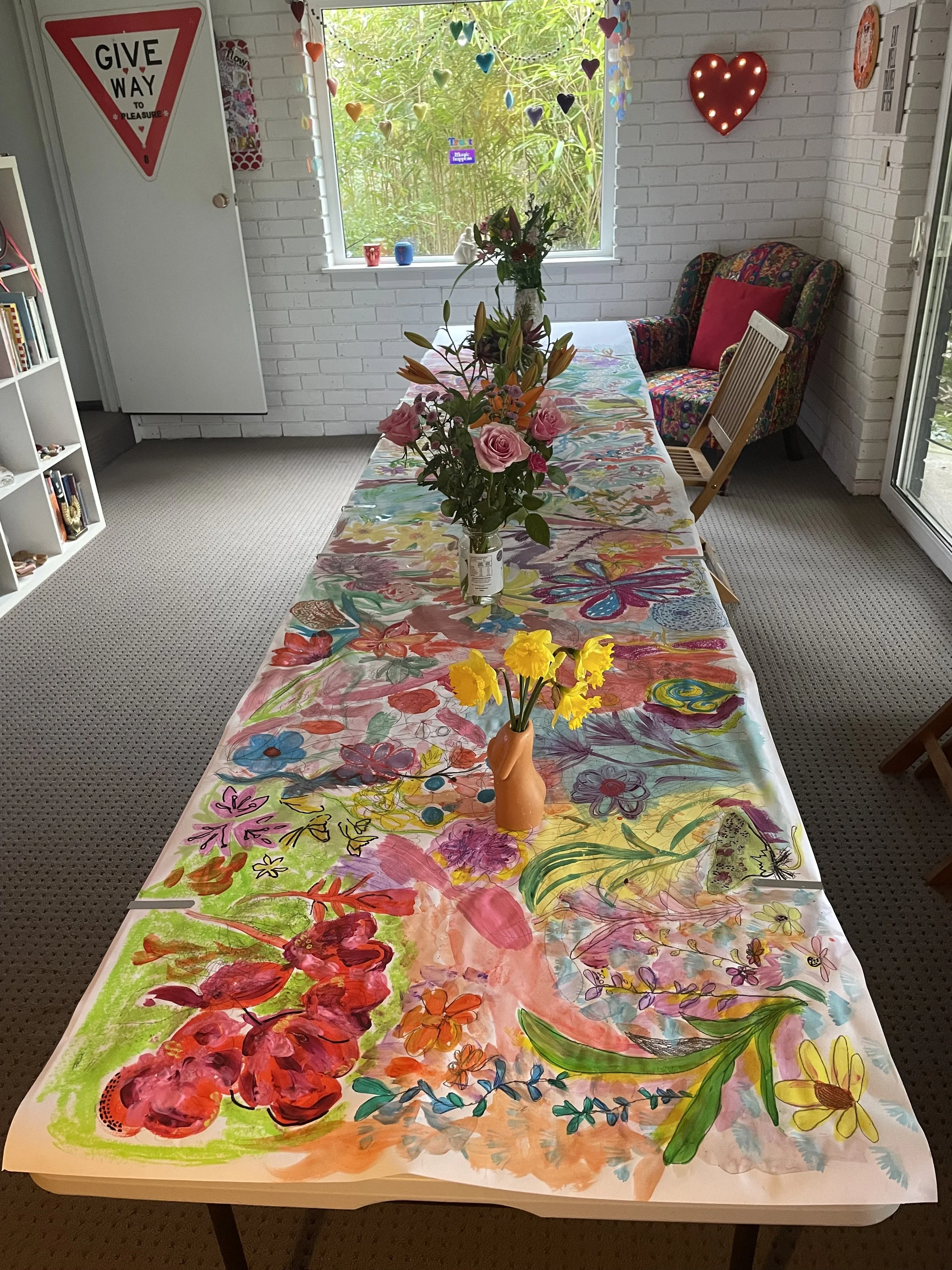 A long table covered with a colorful, hand-painted tablecloth featuring floral and abstract designs. On the table, there is a peach-colored vase with artificial flowers, including pink roses and orange lilies. In the background, a white brick wall ha