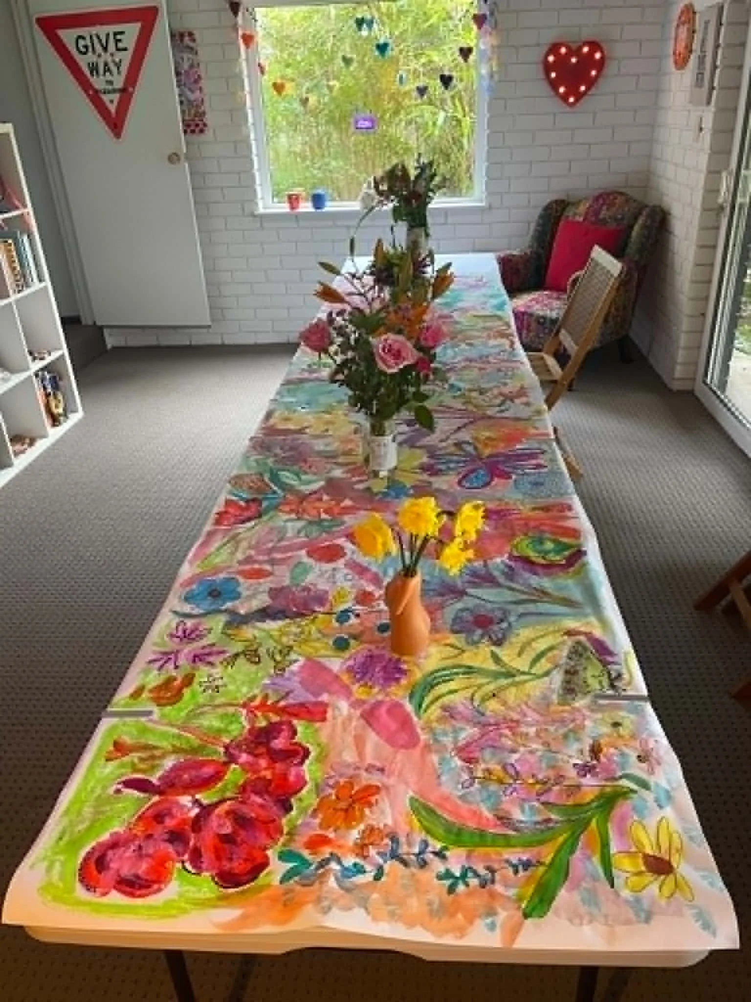 Colorful tablecloth with vibrant watercolor flower designs at a dining table, decorated with a bouquet of pink and orange flowers in a small orange vase, set in a room with white brick walls, minimal furniture, and wall decorations.