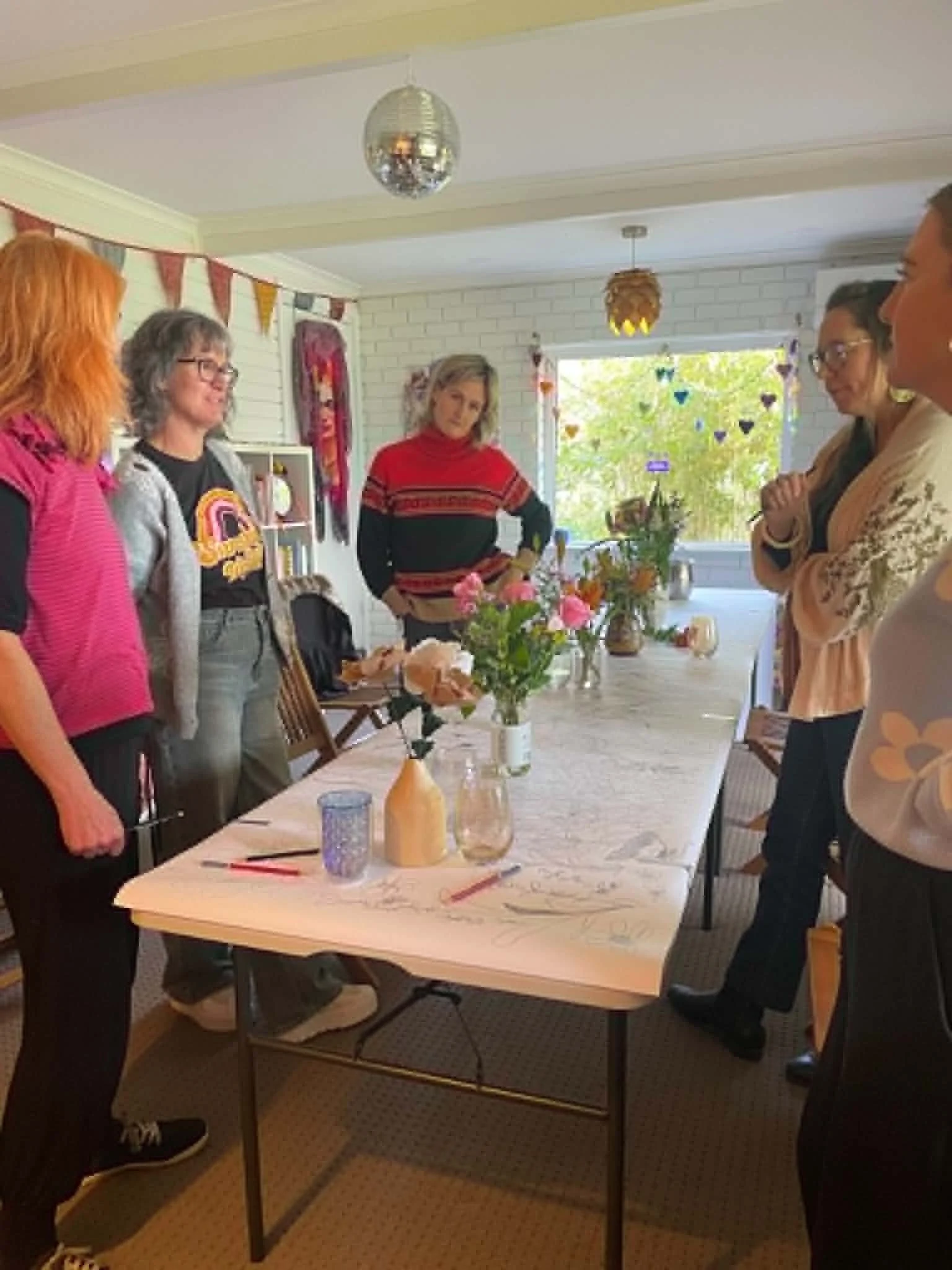 Group of people gathered around a table with flowers, vases, and tableware in a decorated room with bunting and hanging light fixtures.