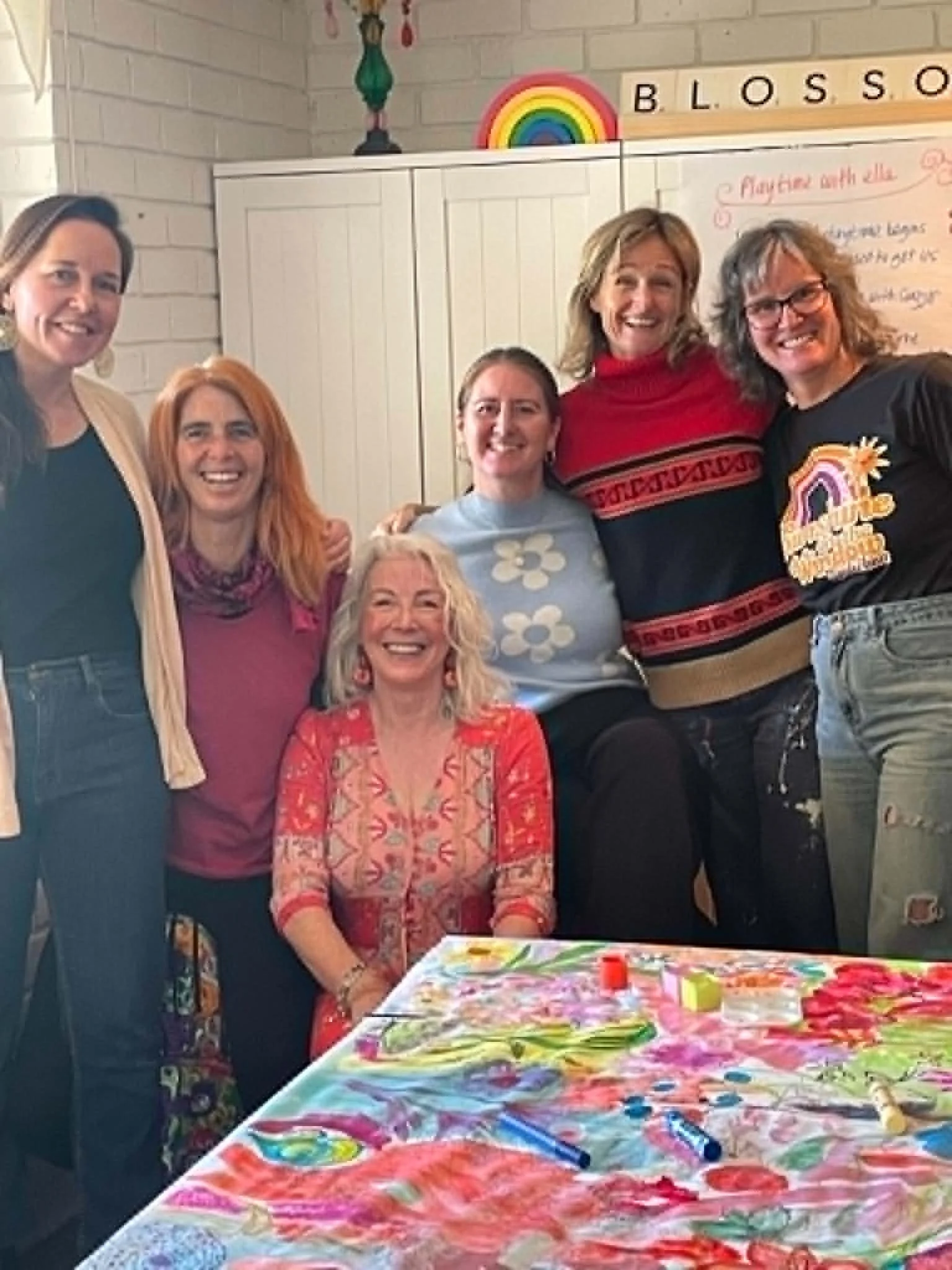 Group of seven people, mostly women, gathered around a colorful art project table in a room decorated with rainbow and flower motifs, smiling and posing for the camera.