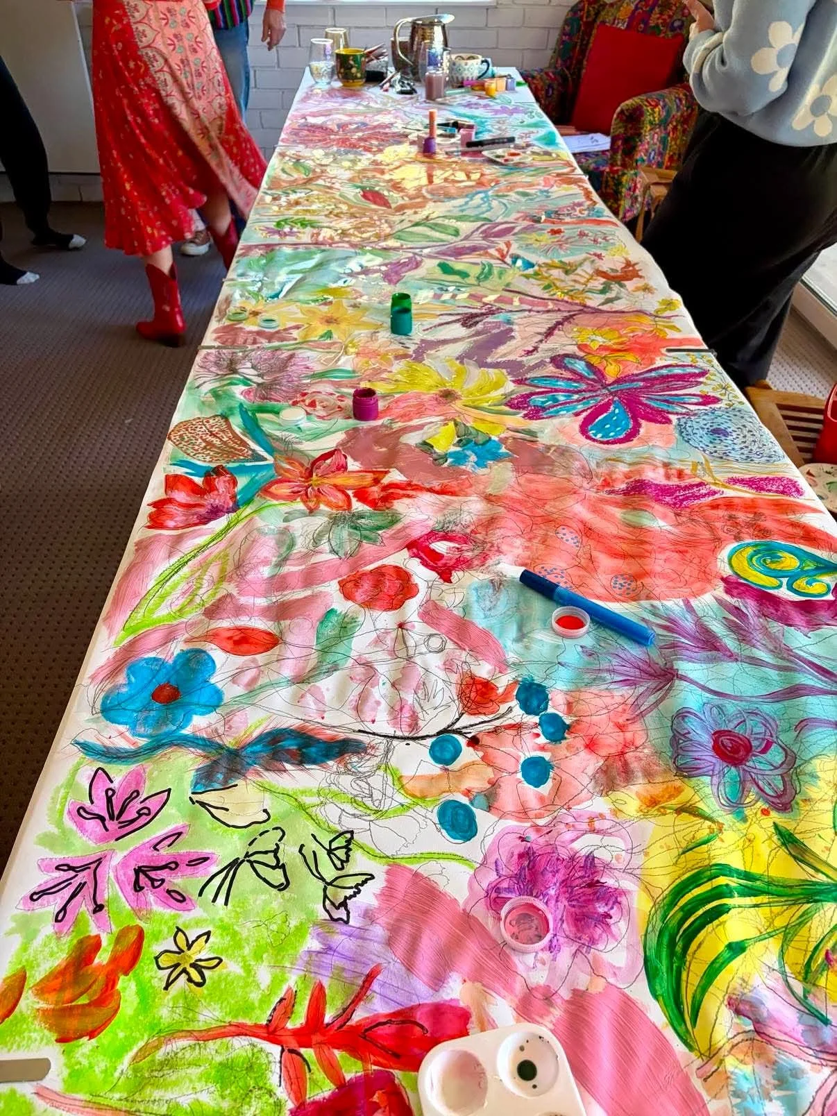 A large table covered with a colorful, hand-painted floral and butterfly design on a generous sheet of paper. Art supplies like markers, paint, and ink bottles are scattered across the table. Several people stand around the table, engaged in creative