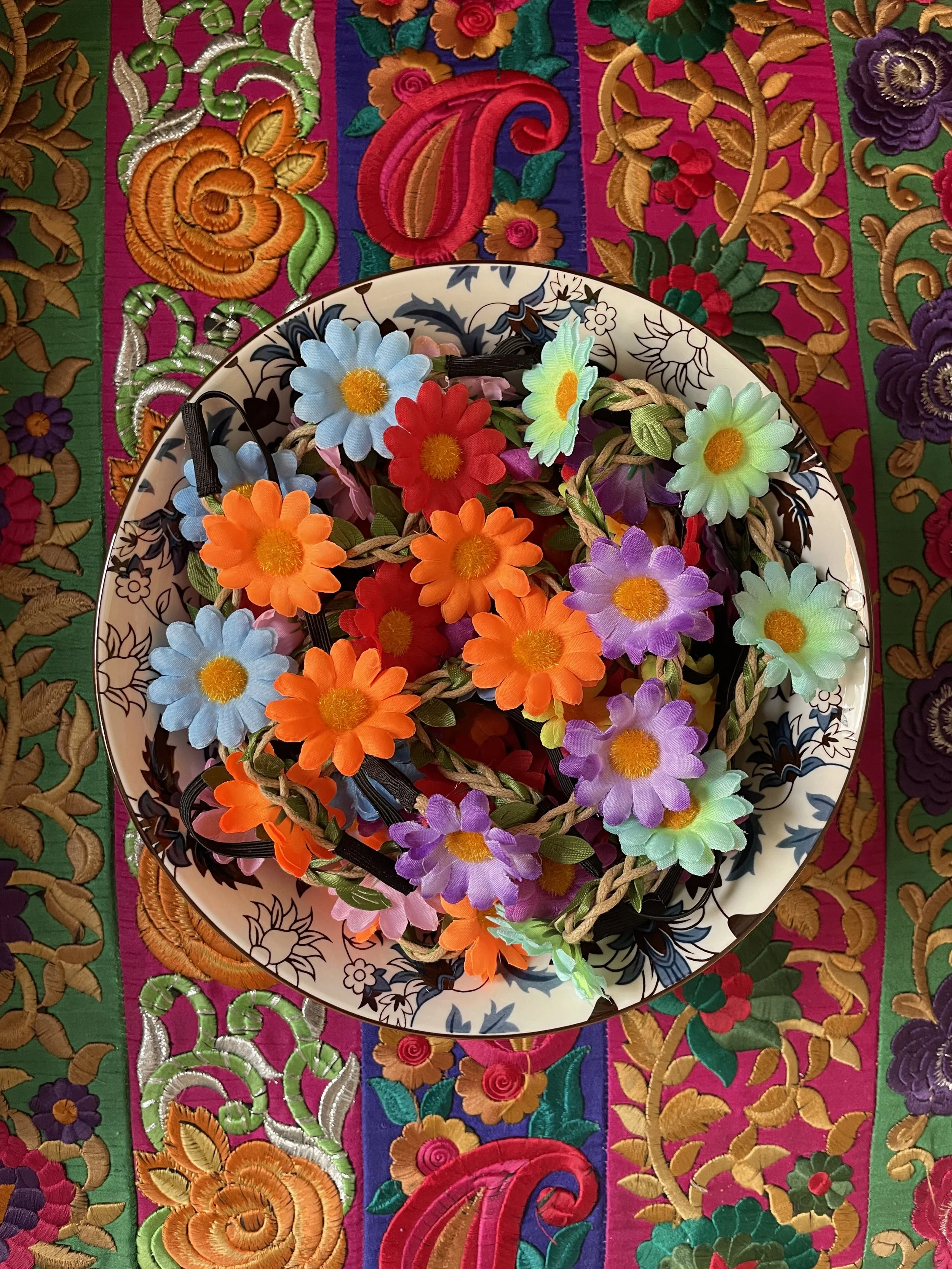 A colorful, embroidered textile background with floral patterns and a tableware plate filled with artificial flower garlands.