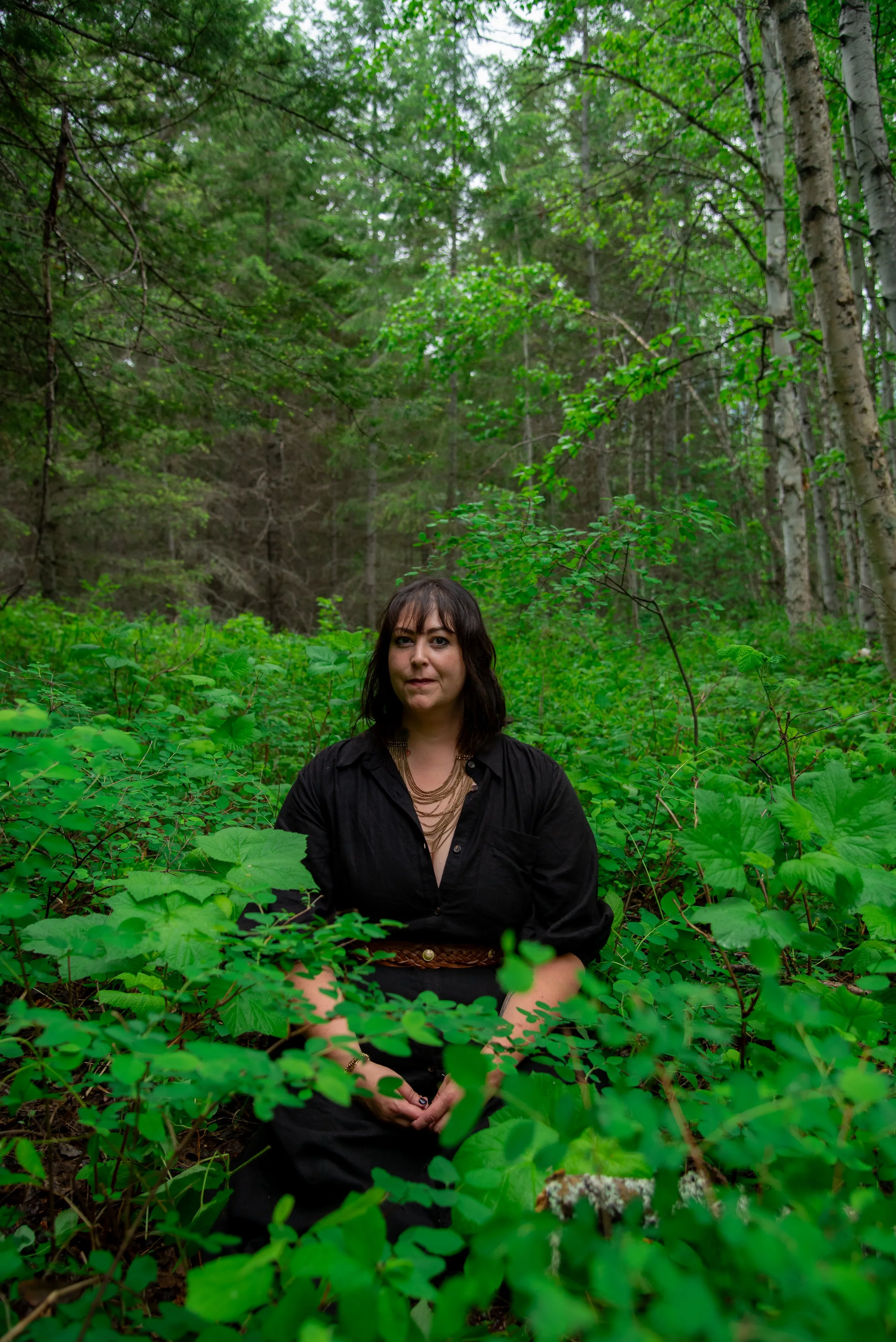 A woman with dark hair and a black shirt sitting among green plants in a forest.