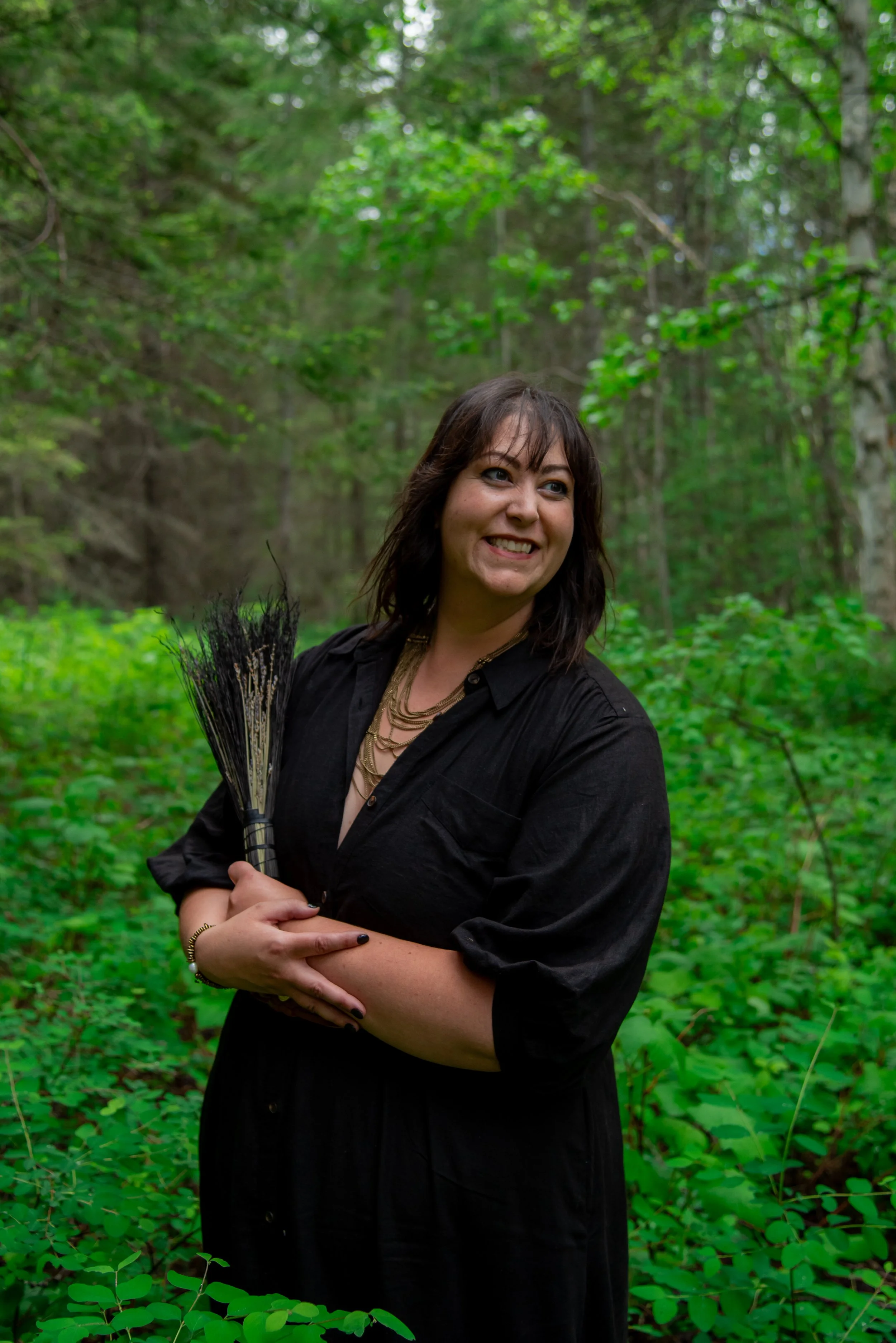 A woman with brown hair and a black outfit holding a bundle of black sticks or flowers, smiling in a lush, green forest.