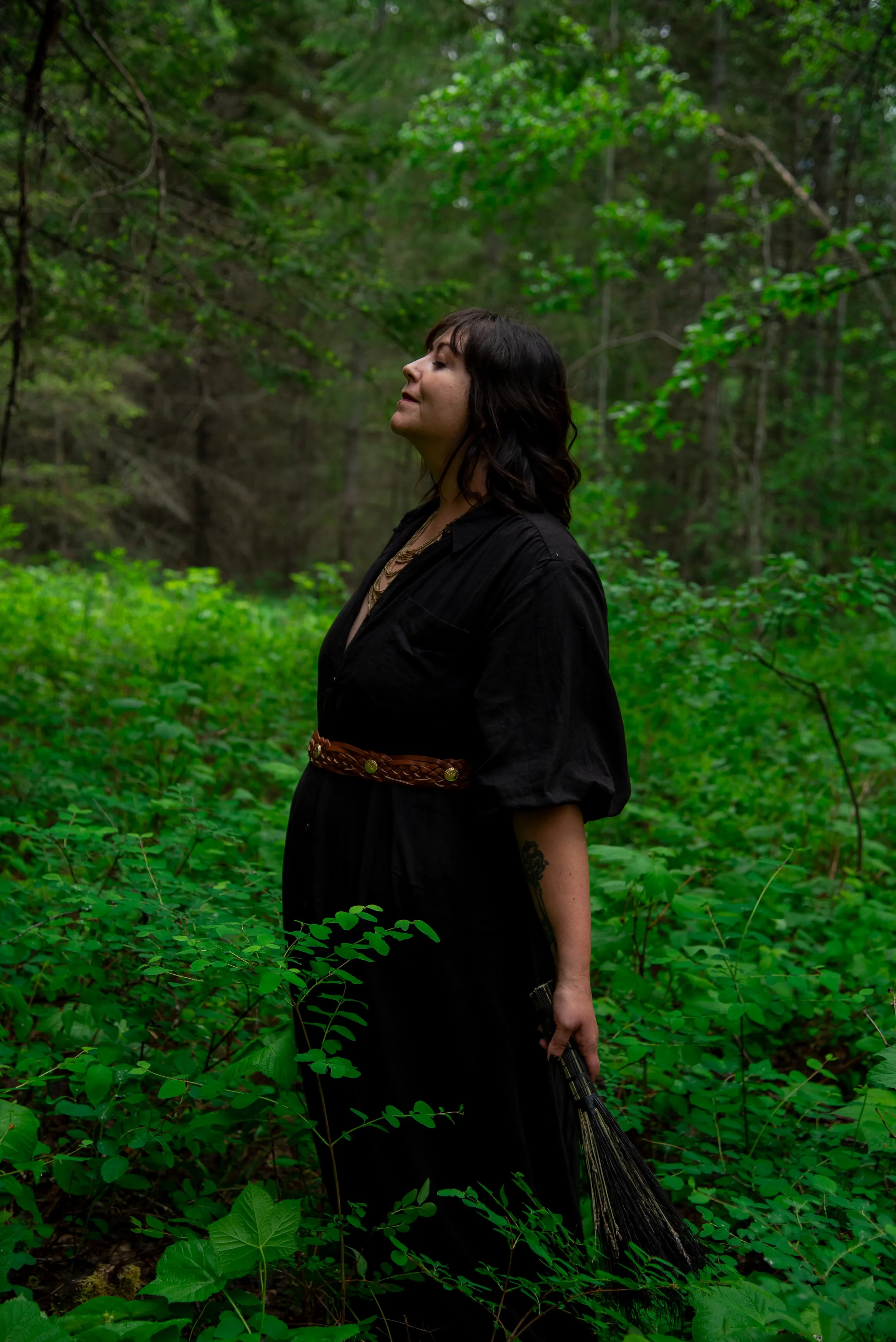 A woman with dark hair and a black dress standing in a lush green forest, holding a fan of herbs in her right hand and appearing to enjoy the peaceful nature around her.