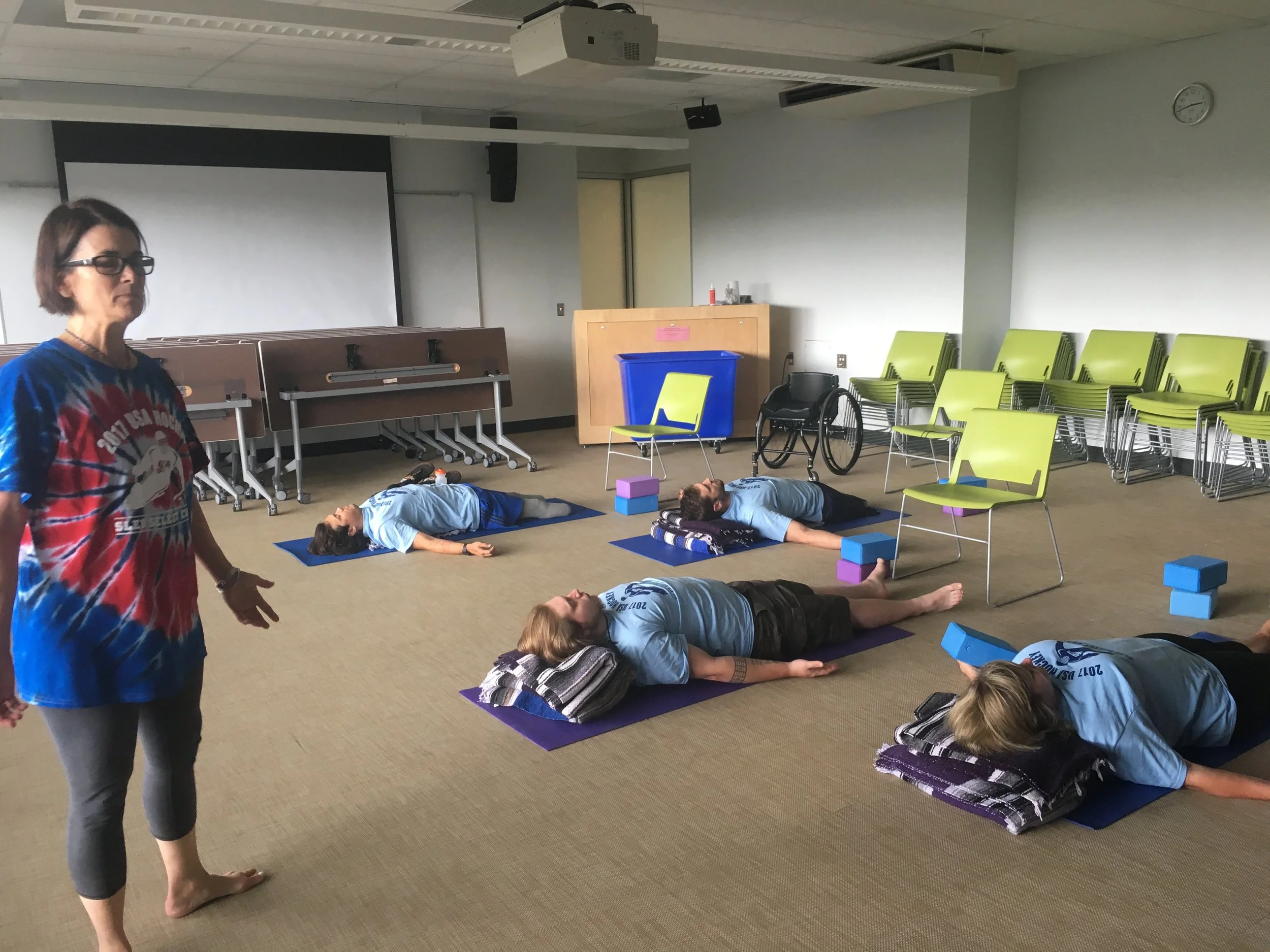 Dr. Sue in a tie-dye shirt standing beside students resting on yoga mats in a classroom