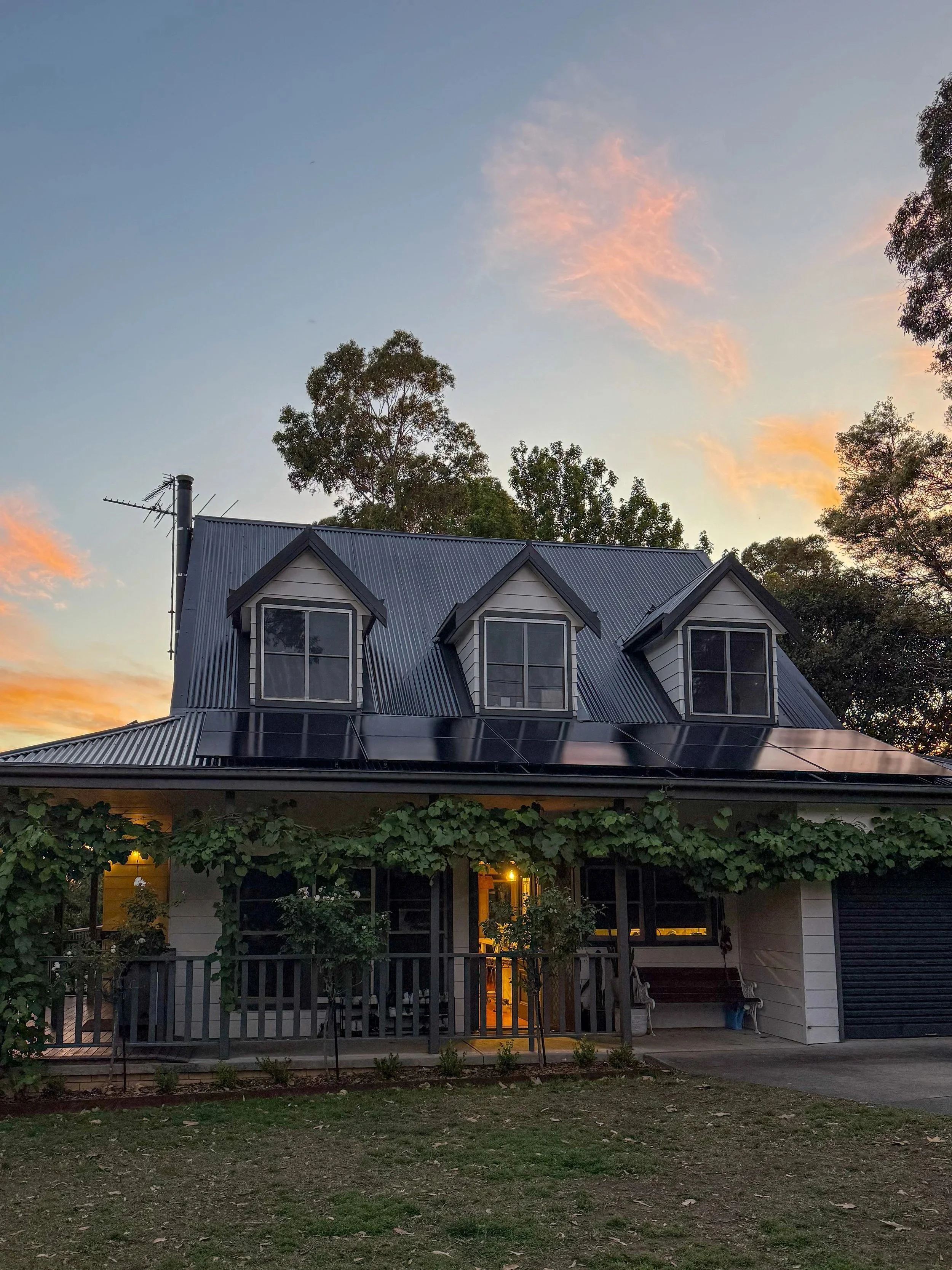 A house with a metal roof, dormer windows, and surrounded by greenery, shown at dusk with a sky filled with pink and orange clouds.