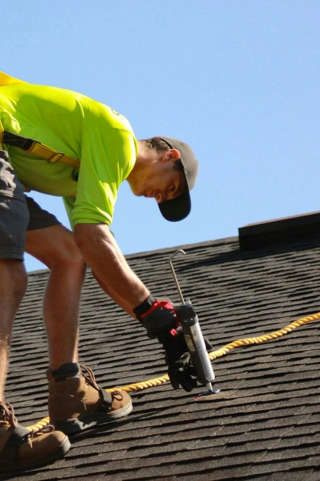 A man wearing a high-visibility yellow shirt, gray shorts, a gray cap, and gloves working on a roof. He is using a caulking gun with a yellow rope on the shingles under bright sunlight.