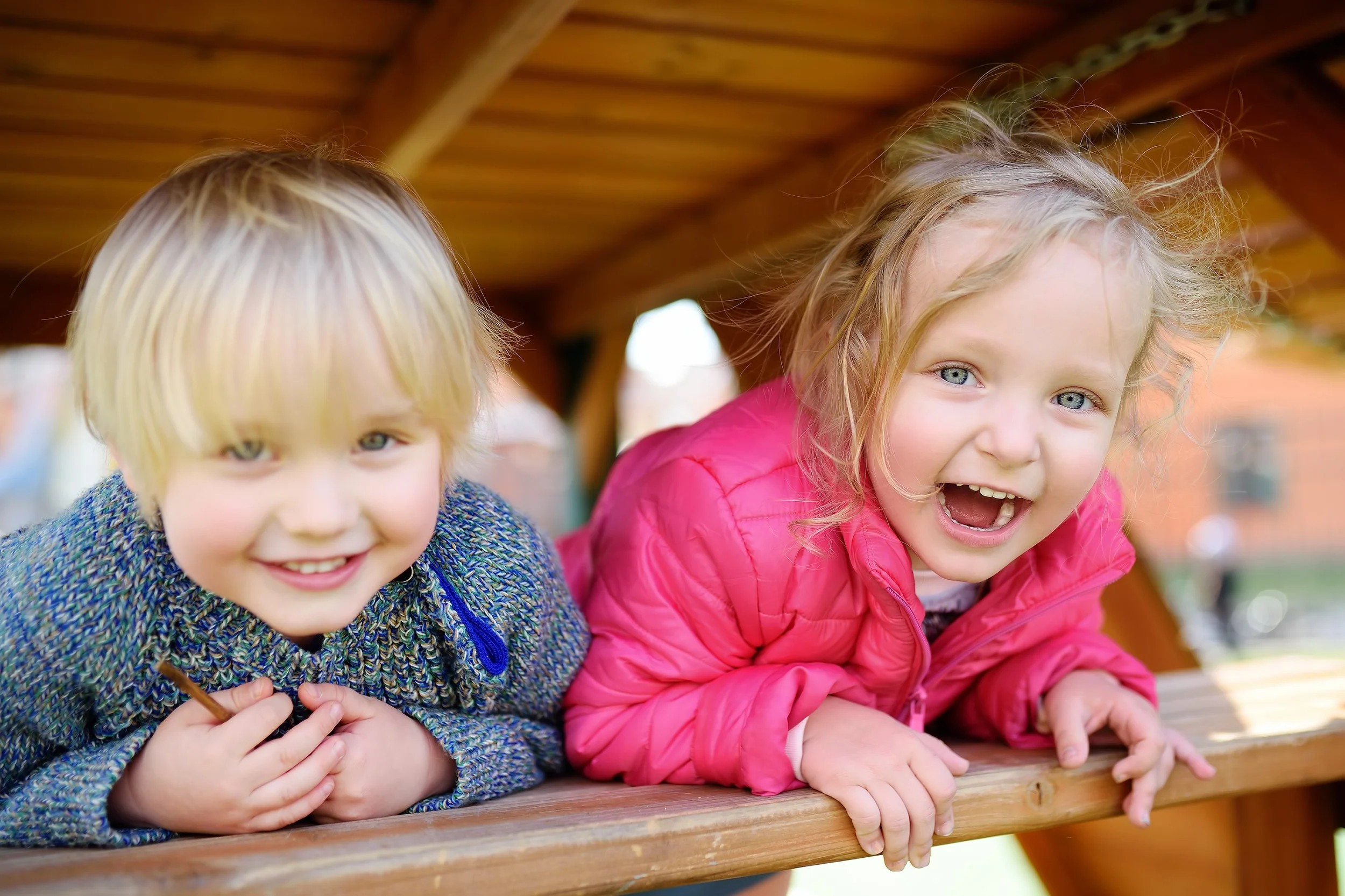 Two young children, a blonde boy and a girl with curly hair, smiling and leaning on a wooden railing outdoors.