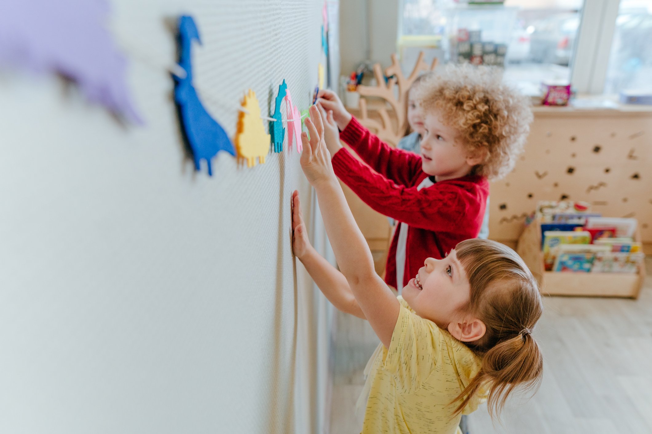 Children decorating a wall with colorful paper cutouts in a classroom.