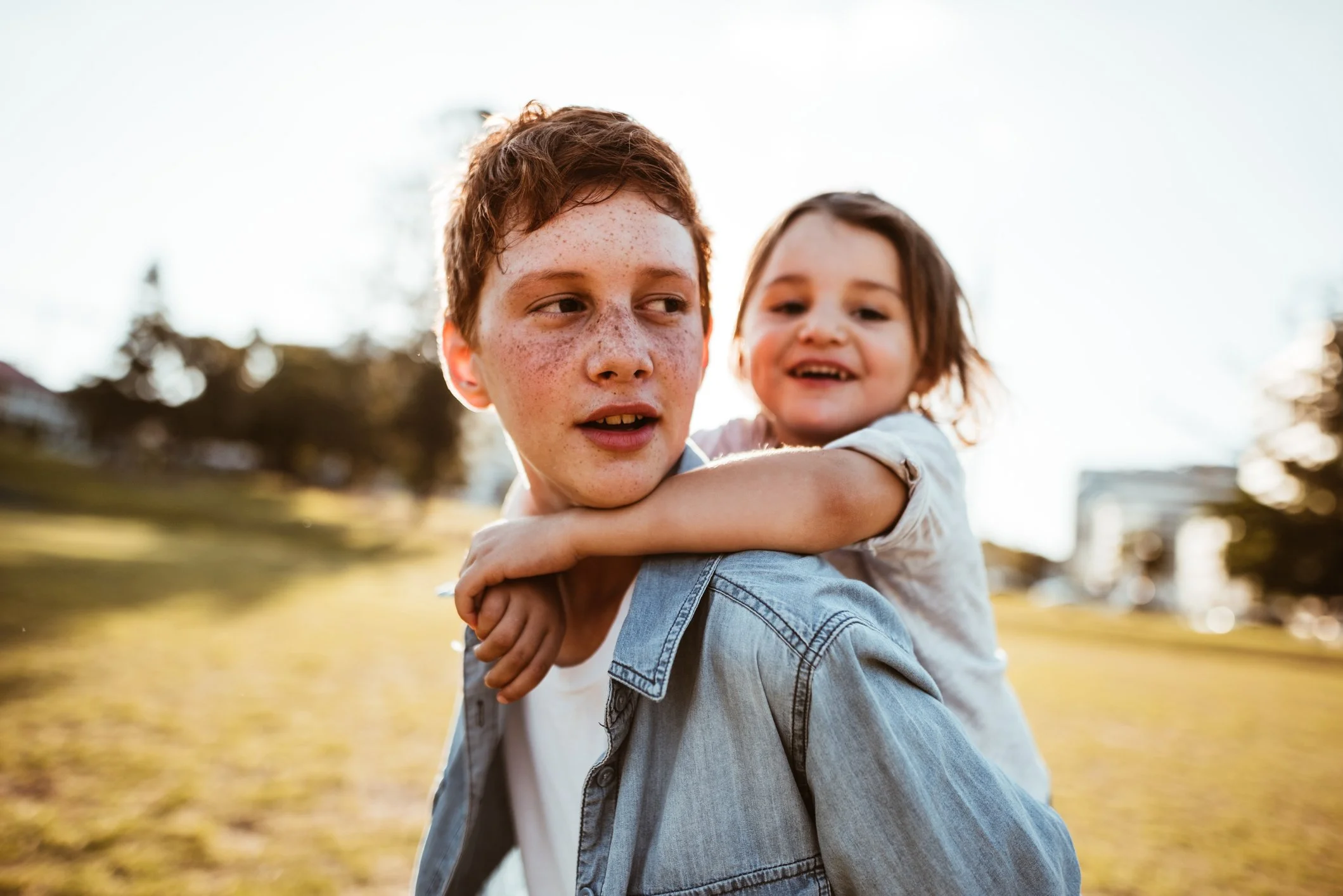 A boy with red hair and freckles carrying a young girl on his back outdoors in a park during sunset, with trees in the background.