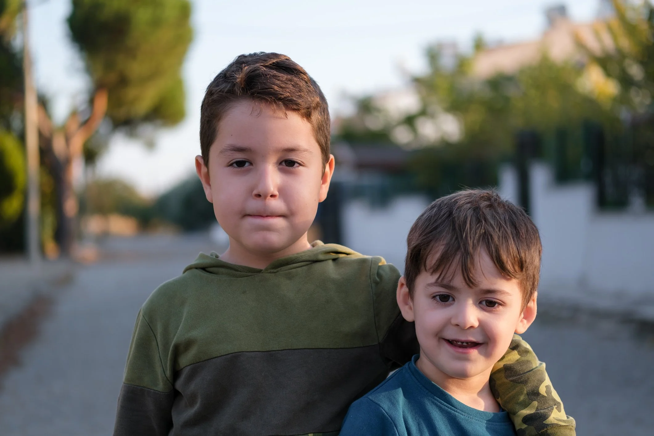 Two young boys standing outdoors in a park or street, with trees and buildings in the background. The older boy has short brown hair and is wearing a green and gray hoodie, looking directly at the camera with a neutral expression. The younger boy, partially visible, has brown hair and is wearing a blue shirt with camouflage sleeves, smiling with his teeth showing.