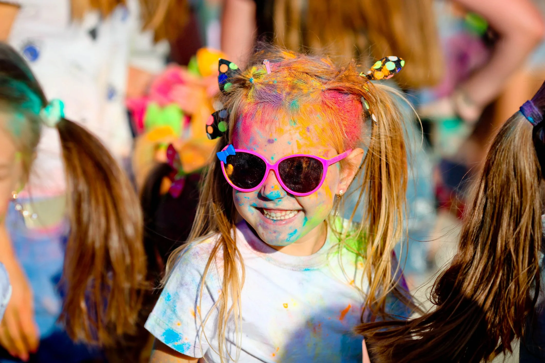 A young girl with red hair wearing pink sunglasses, smiling, covered in colorful paint with other children participating in a messy outdoor event.