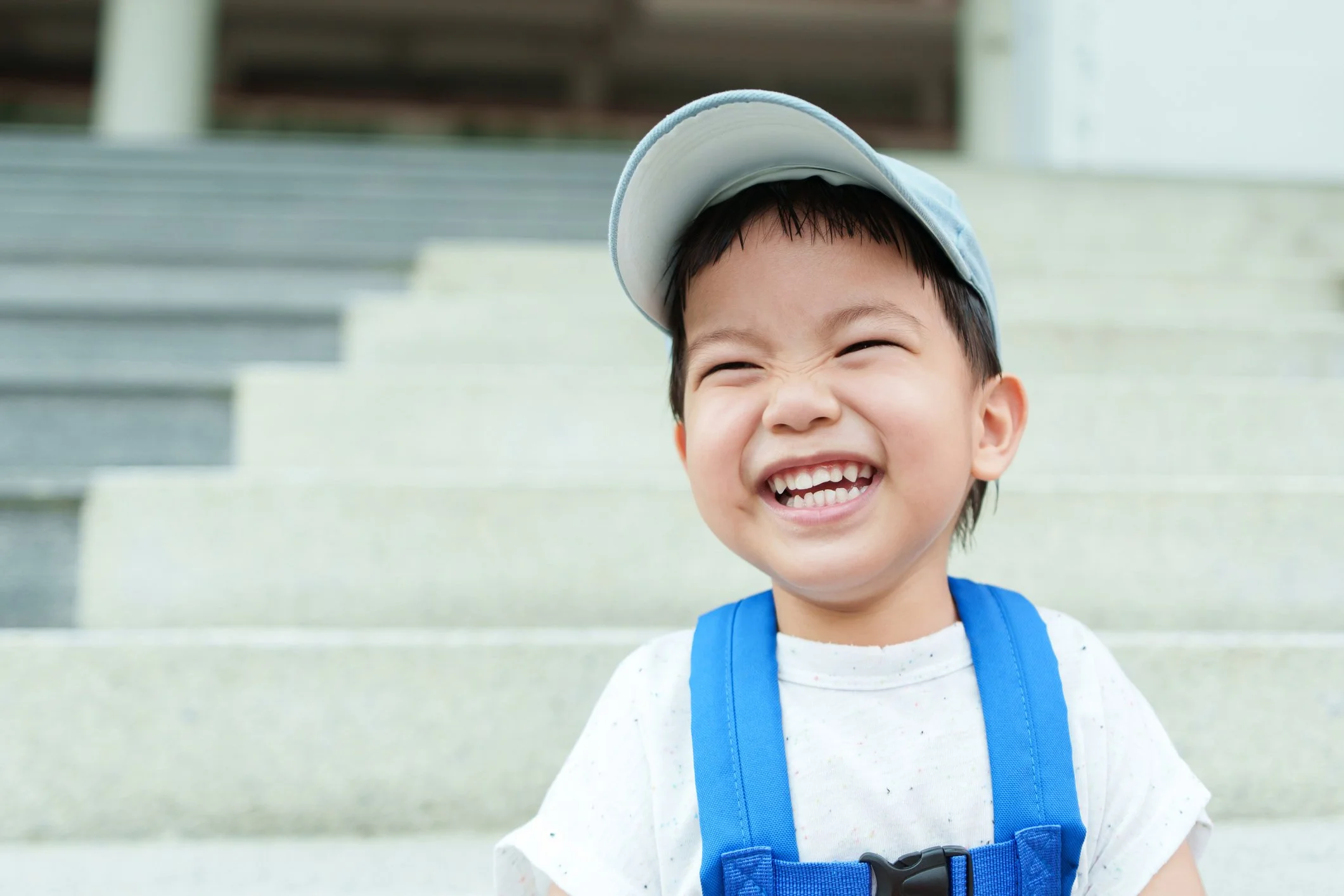 A young boy wearing a white t-shirt with colorful specks, a blue backpack, and a light blue cap, smiling broadly with eyes squinted, standing outdoors in front of concrete steps.