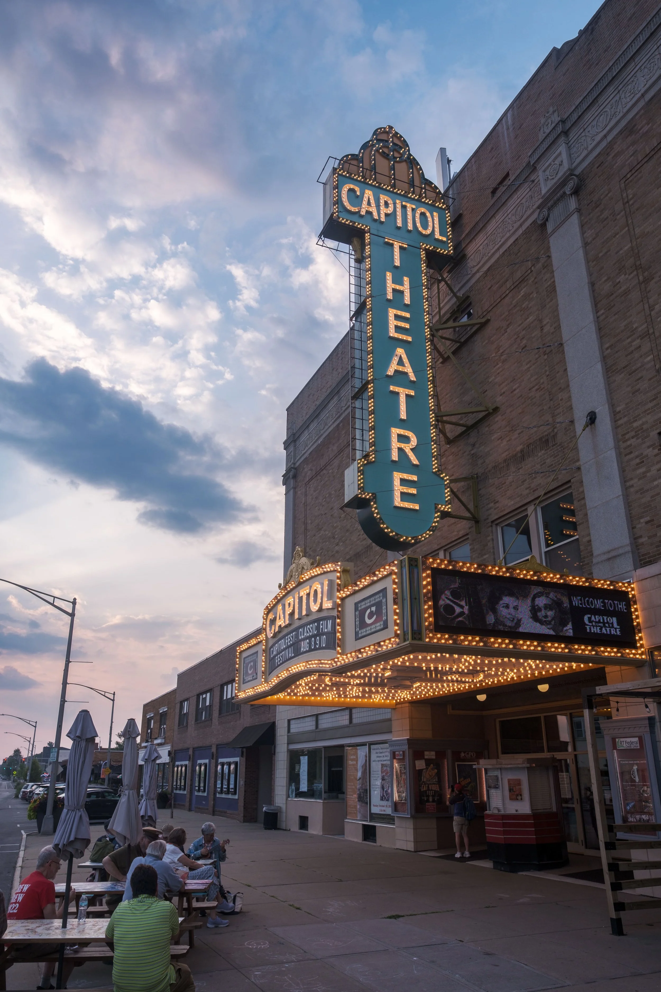 People sitting at outdoor tables in front of a theater with a large illuminated sign reading 'Capitol Theatre' and a marquee advertising a film festival, with a brick building and a cloudy sky in the background.