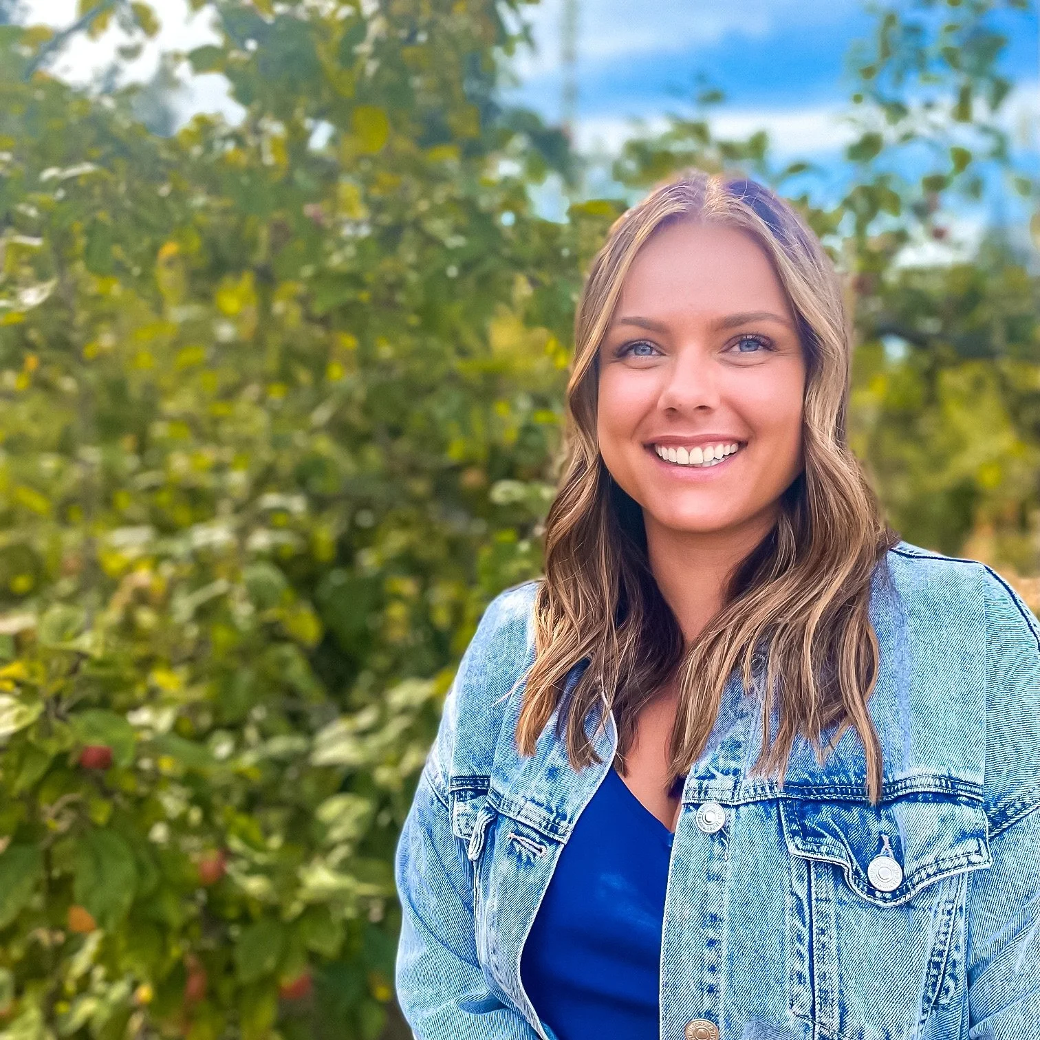 A smiling young woman with long, wavy blonde hair and blue eyes, wearing a denim jacket over a blue top, standing outdoors with green leafy trees in the background.