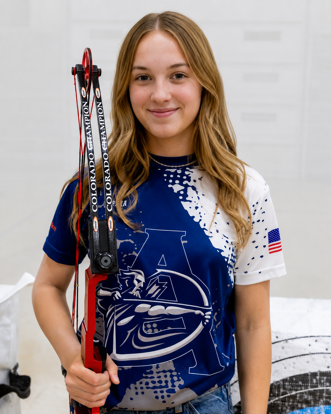Young woman with long wavy hair holding a pair of colored archery bows, wearing a blue and white sports jersey with an American flag patch on the sleeve, smiling at the camera.