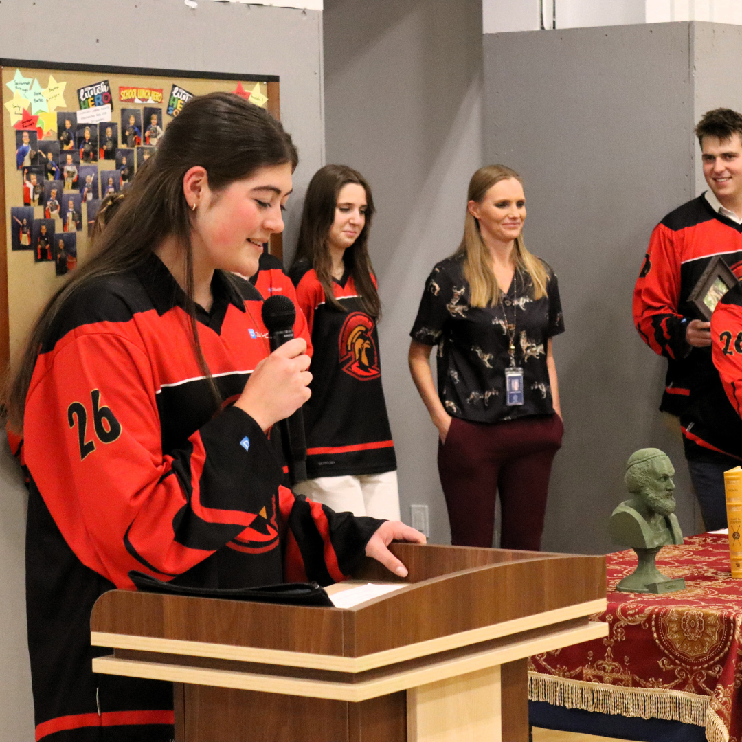 A young woman in a red and black hockey jersey stands at a wooden podium, speaking into a microphone, with a small bust sculpture and a yellow container on a table nearby. Four other women and men in similar hockey jerseys and a woman in a black blouse with a lizard print are standing behind her in an indoor setting.