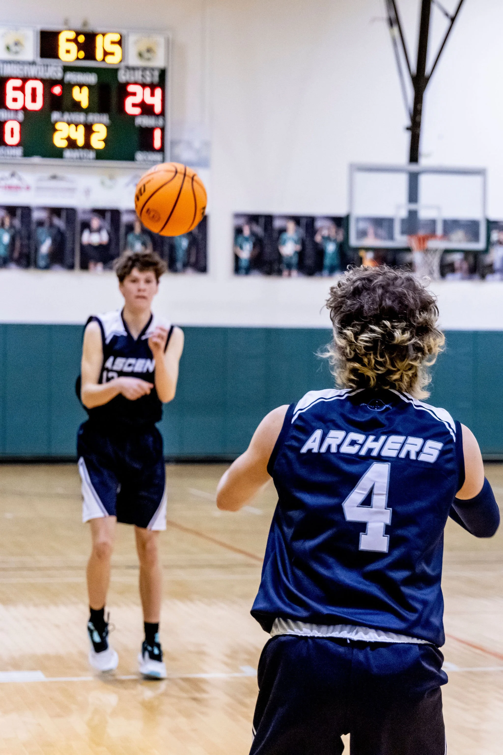 Two young boys playing basketball indoors. The boy in the foreground wears a dark blue jersey with the number 4 and the word 'ARCHERS' on the back. The boy in the background is preparing to catch or pass the ball. The scoreboard shows 6:15 remaining, with the visiting team leading 60-24 in the fourth quarter.
