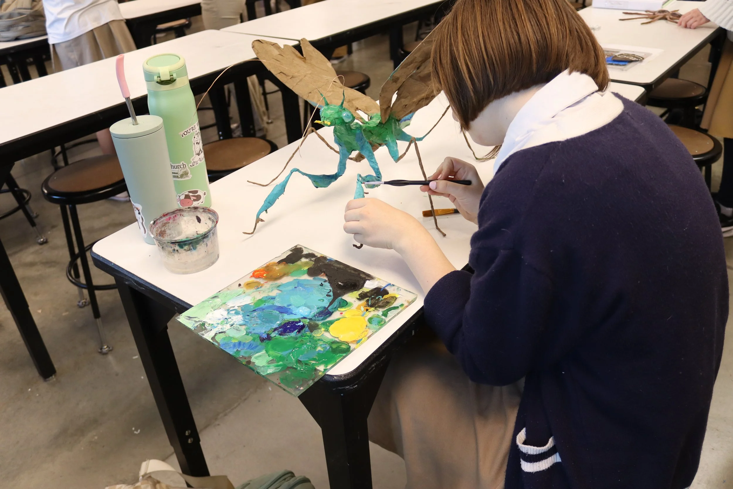A person with short brown hair, wearing a white hoodie and dark sweater, painting a 3D model of a dragon with green body and brown wings on a table covered with art supplies in a classroom or studio.