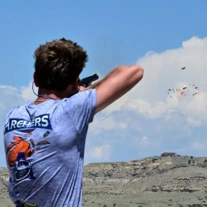 A person shooting a firearm outdoors under a blue sky with clouds, with a landscape and hills in the background.
