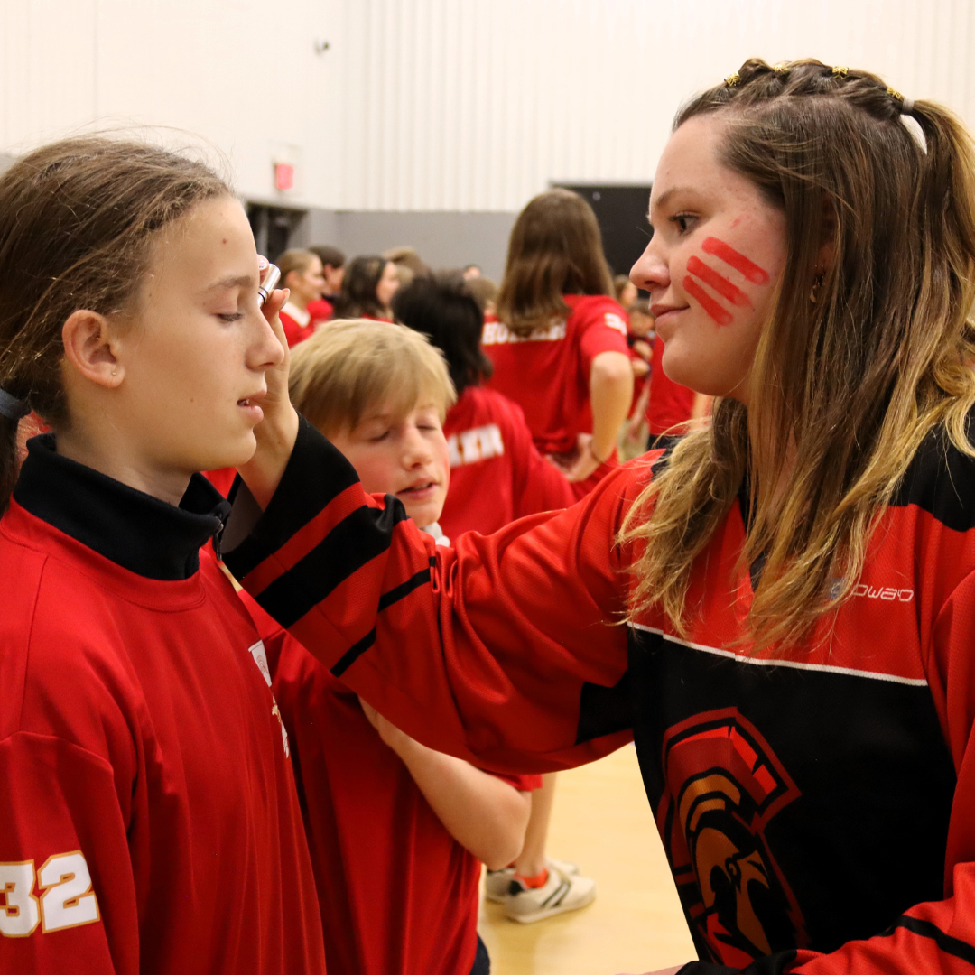 A young girl with face paint of red stripes is gently placing her hand on the forehead of another girl, who appears to be listening or receiving encouragement, during a team event with many children wearing red sports jerseys.