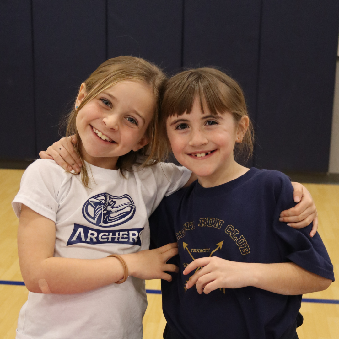 Two young girls smiling and hugging each other indoors, one wearing a white T-shirt with an archer logo and the other wearing a navy blue T-shirt with a running club logo.