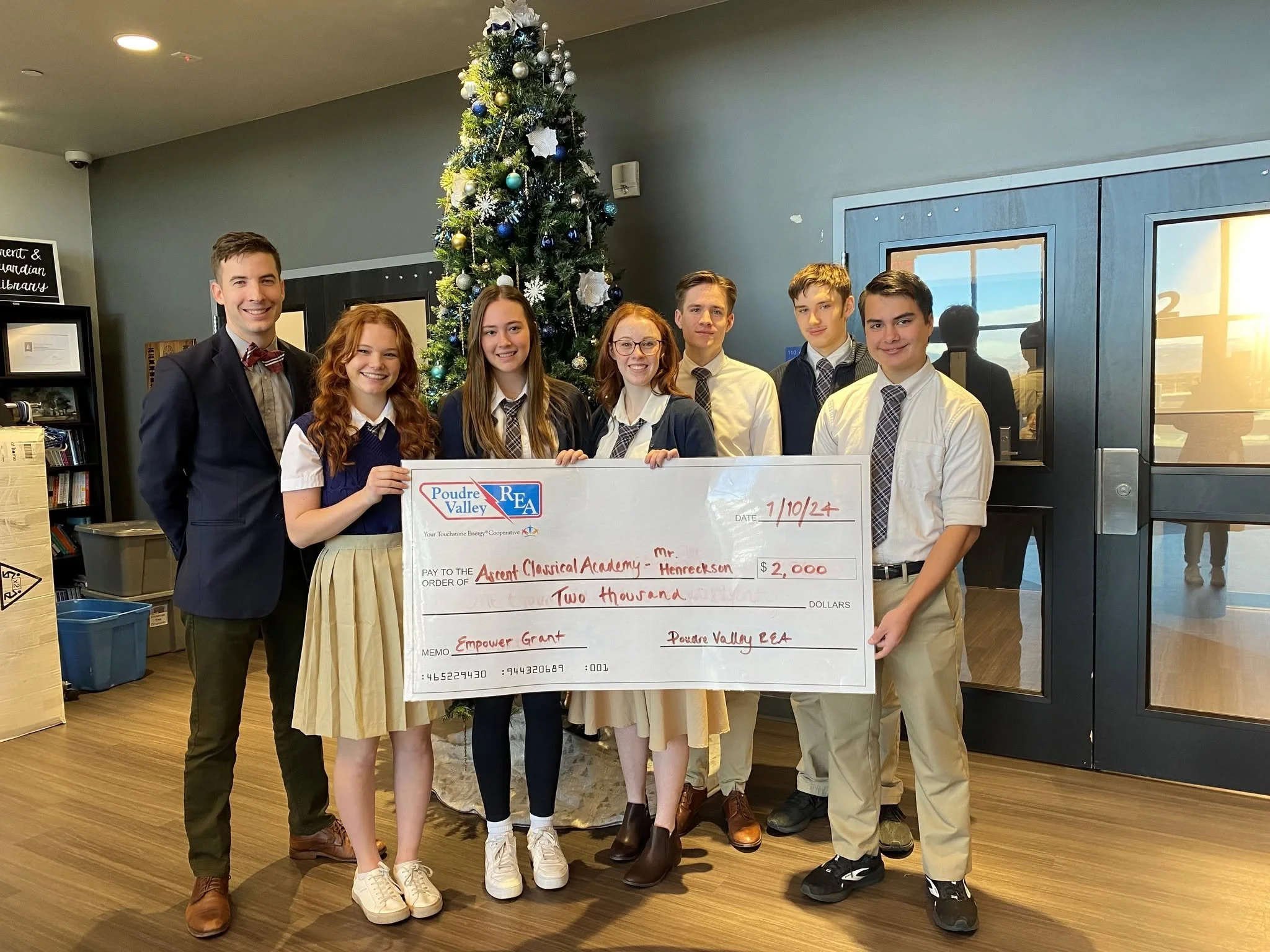 A group of seven young people, dressed in semi-formal attire, is holding a large check. They are standing in a room decorated with a Christmas tree, which is decorated with ornaments and white ribbons. The group is smiling and standing close together, with some people holding onto the check. The background includes windows, a bookshelf, and a sign that reads 'Parent & Guardian Library'.