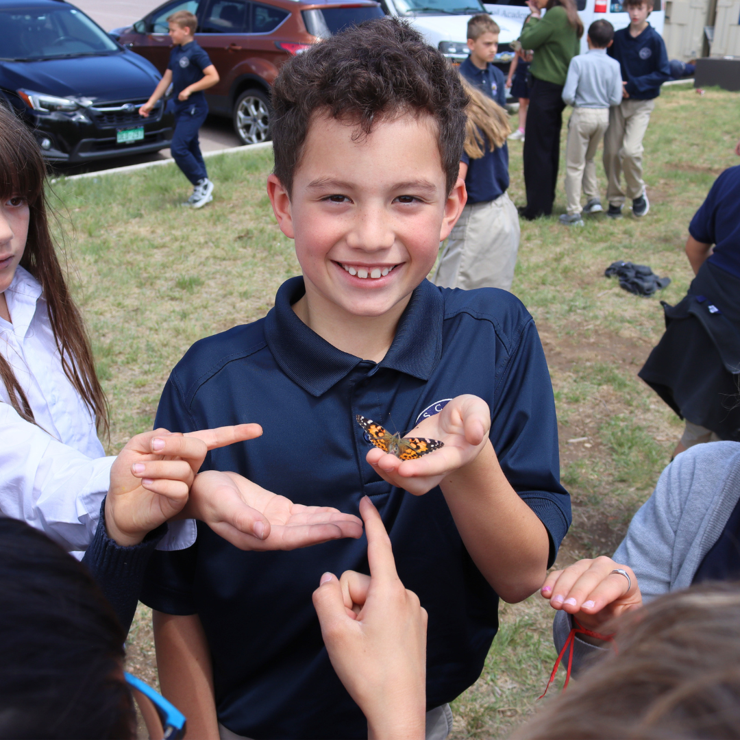 A smiling boy holding a butterfly in his hand surrounded by children outdoors, with cars and people in the background.