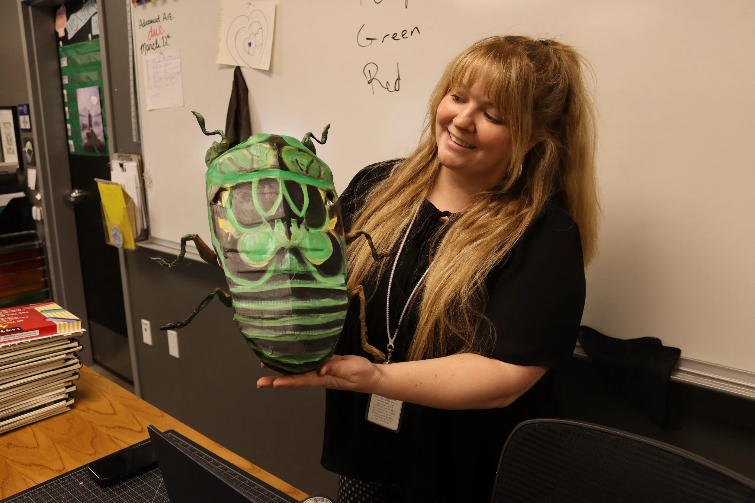 A woman with long, wavy, light brown hair smiling and holding a large, colorful, paper mache or painted model of a bug or beetle in a room with a whiteboard and roller shelves in the background.