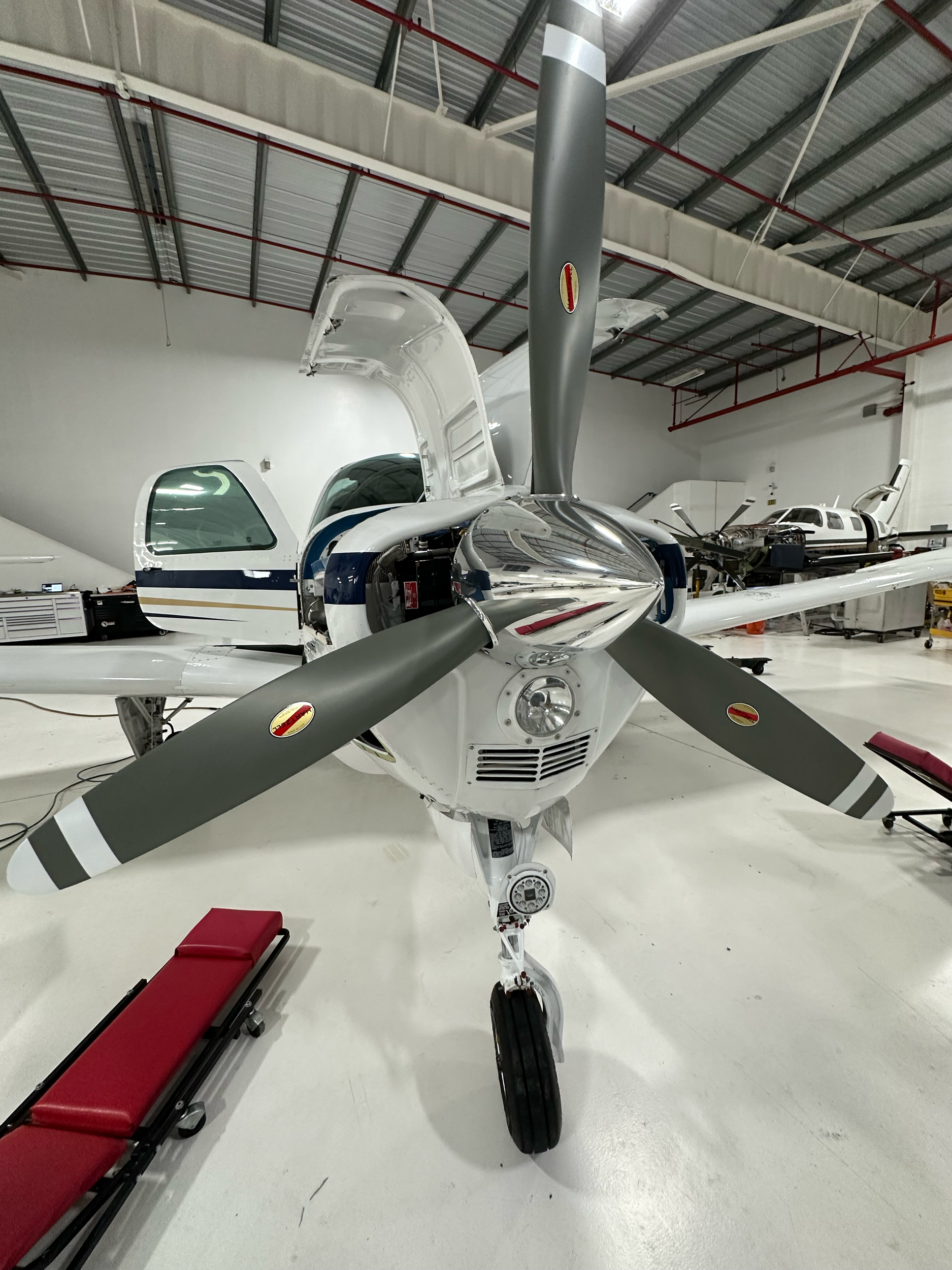Indoor airplane hangar with a small single-engine aircraft in the foreground, surrounded by other planes and maintenance equipment.