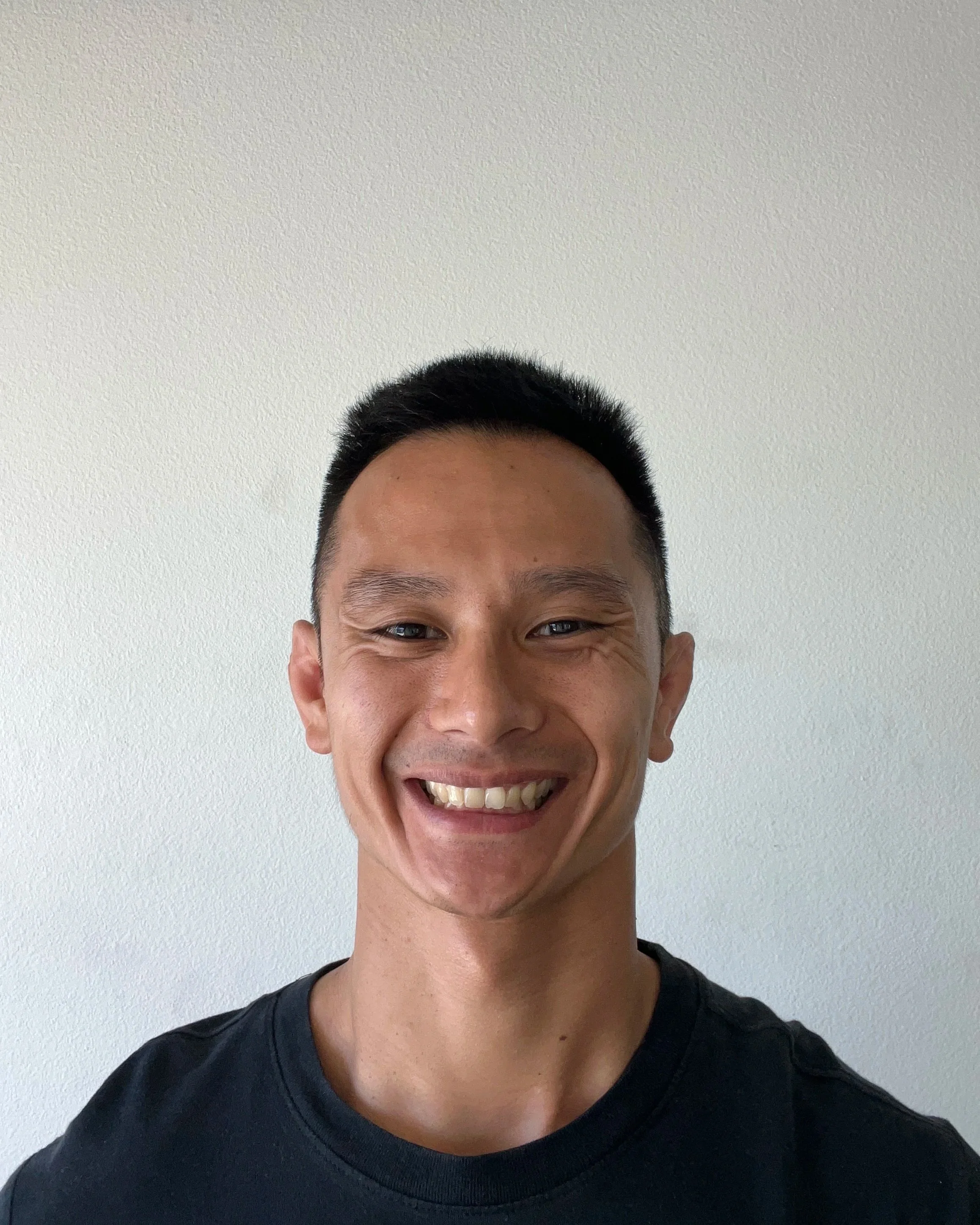 Smiling man with short black hair, wearing a black T-shirt, standing against a plain white wall.