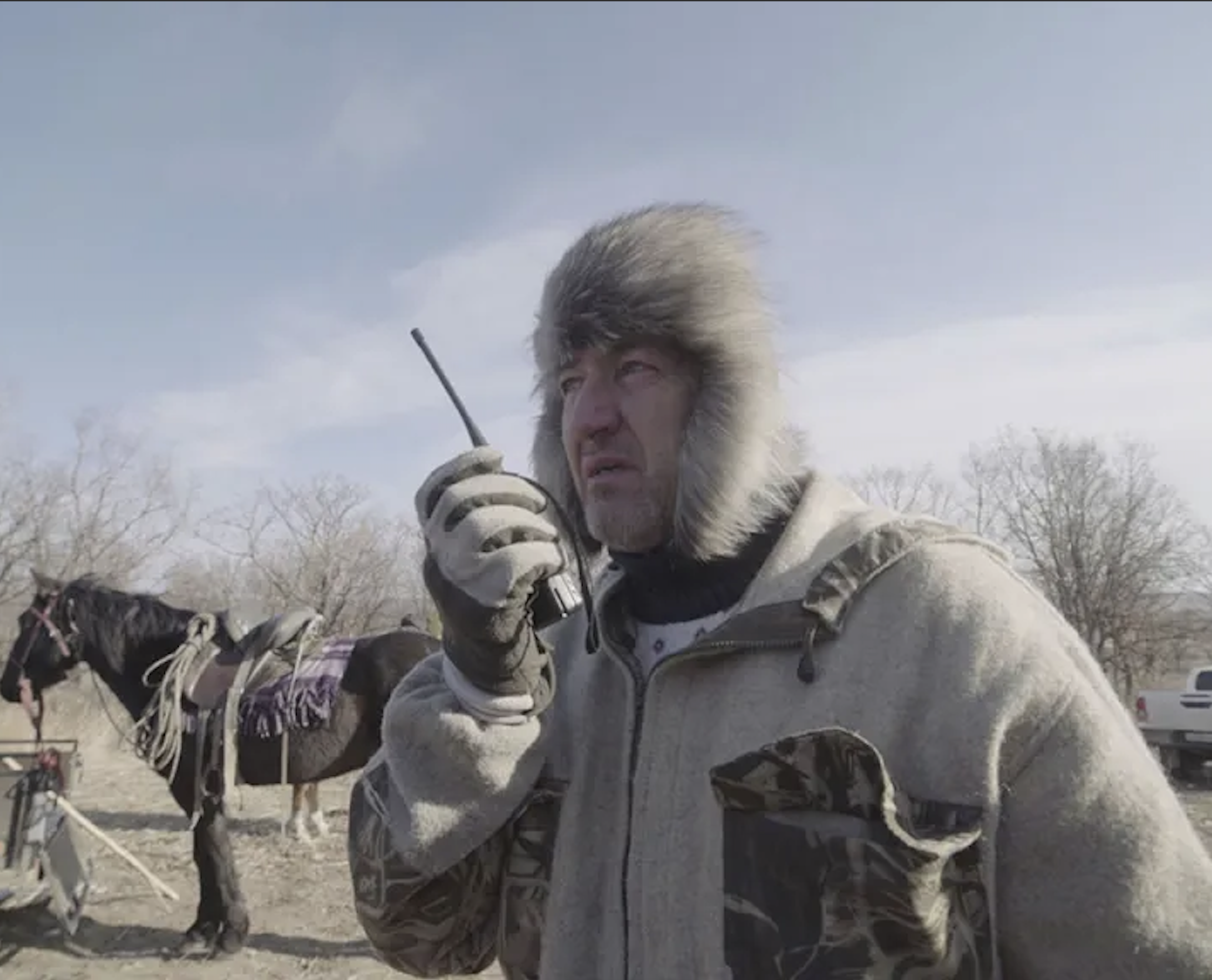 A man wearing a heavy winter coat with a fur-lined hood and gloves holding a radio or walkie-talkie amid a snowy outdoor area with leafless trees and horse-drawn carriage in the background.