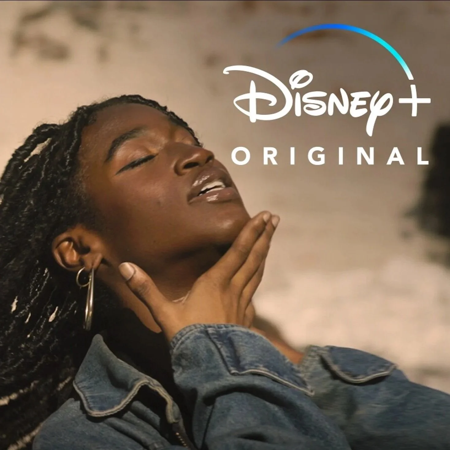 A woman with braided hair and hoop earrings, wearing a denim jacket, holding her chin with her hand, with her eyes closed, as the Disney+ Original logo overlaps the image.
