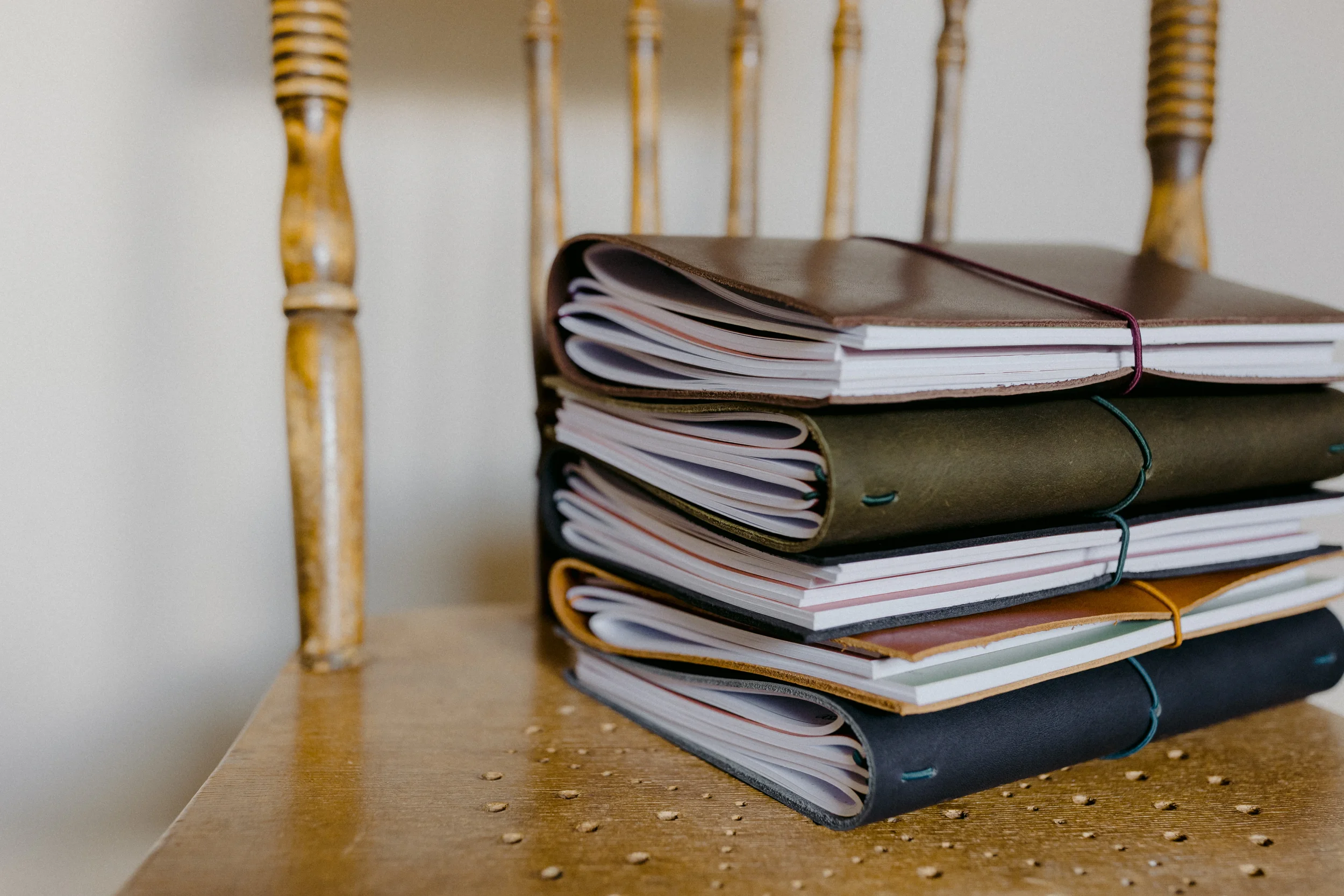 Stacked notebooks and binders on a wooden surface with a wooden chair in the background.