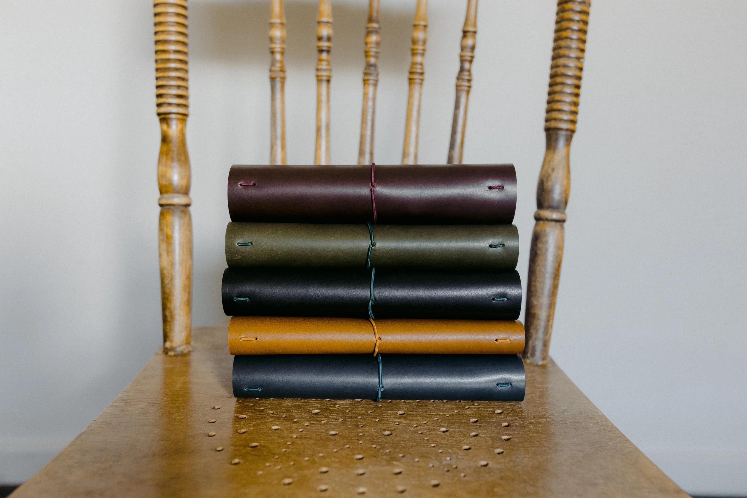 A wooden chair with a spindled backrest and rounded legs, holding five rolled notebooks in yellow, black, gray, green, and maroon, tied with matching colored elastics, on a wooden surface with perforated pattern.