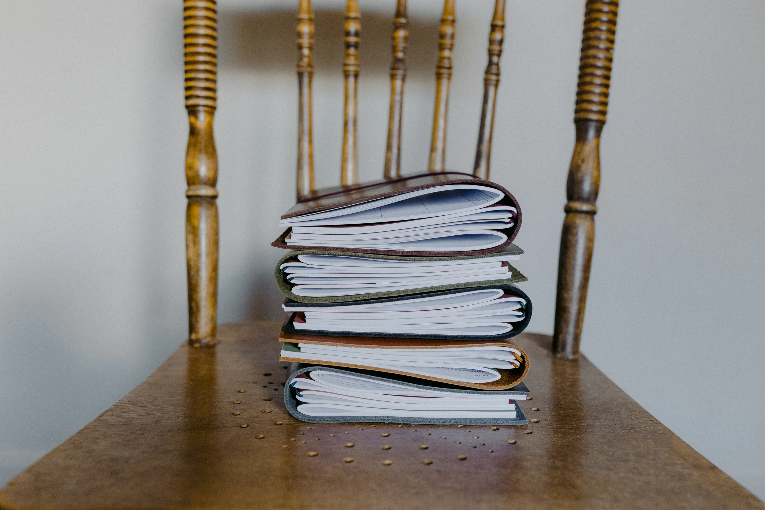 A wooden chair with spindled backrest and armrests behind a small wooden table. On the table, there is a stack of notebooks or journals with various colored covers, some open. The background is a plain wall.