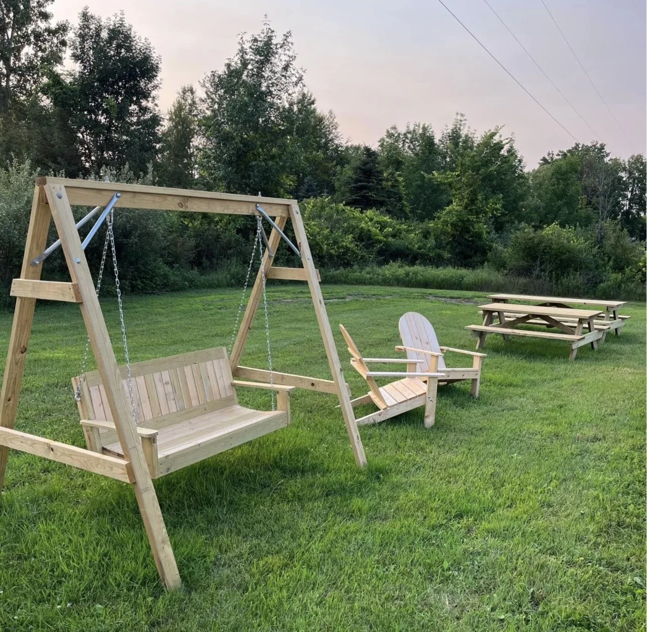 A playground area with a wooden swing set, a wooden Adirondack chair, a wooden bench, and a picnic table on a grassy field surrounded by trees.