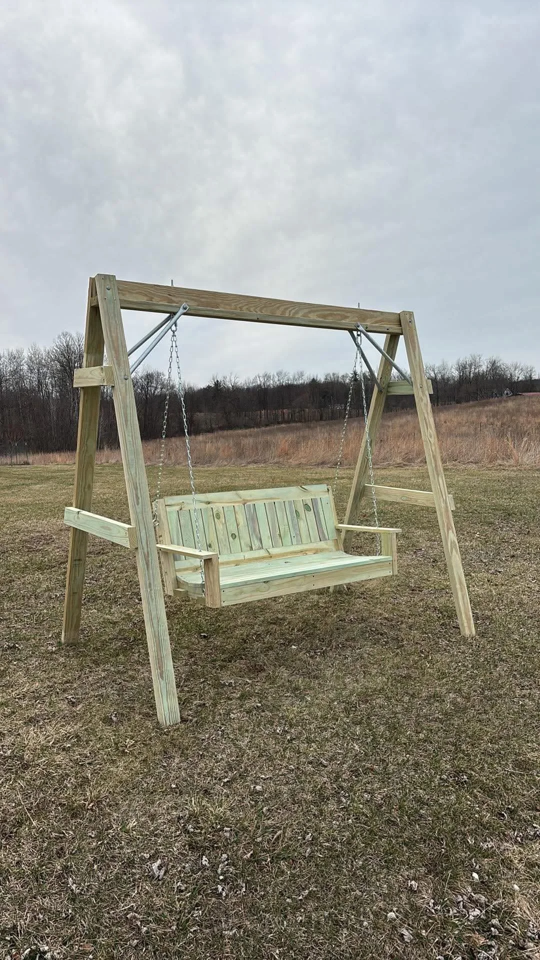 A wooden outdoor swing set with a bench swing in an open grassy field under a cloudy sky.