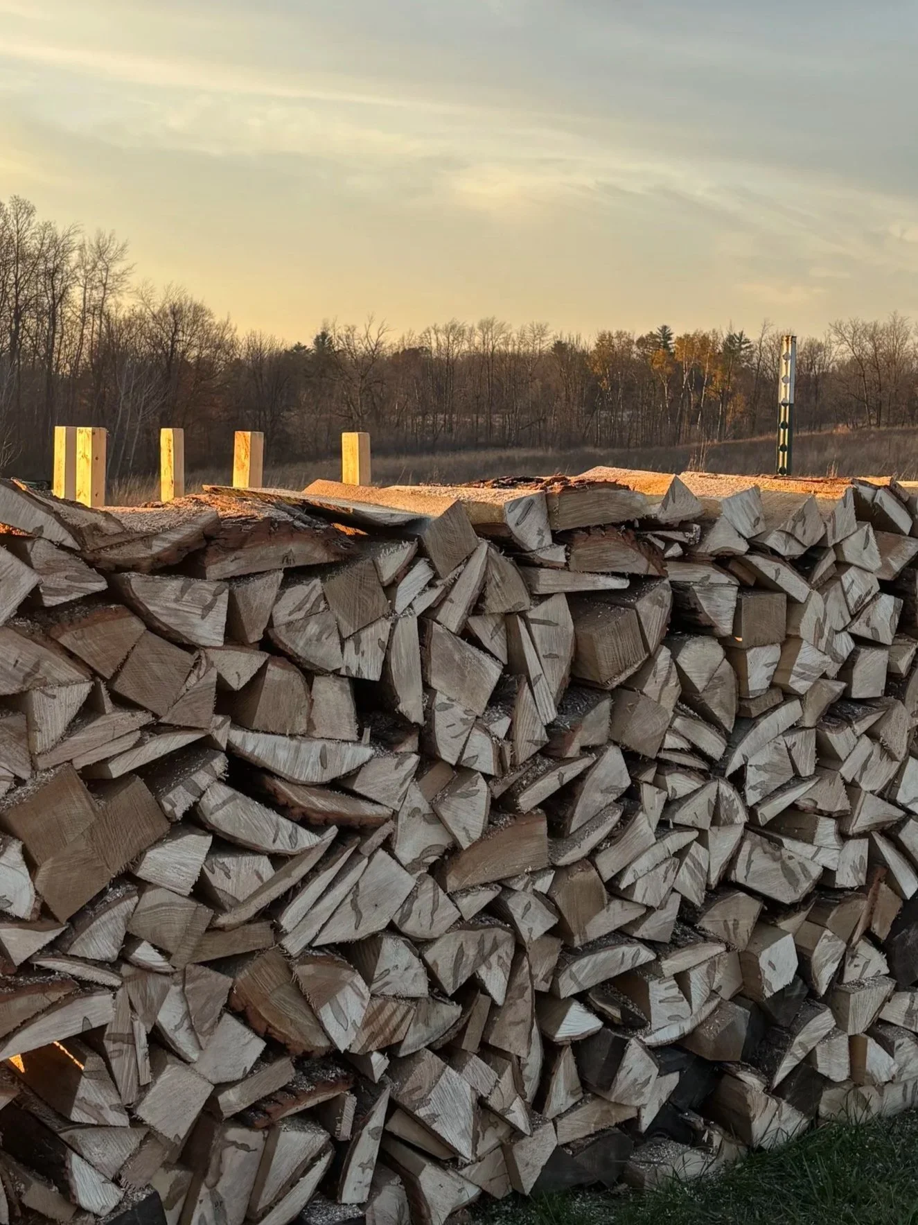A large stack of firewood neatly arranged outdoors, with a background of trees and a partly cloudy sky at sunset.