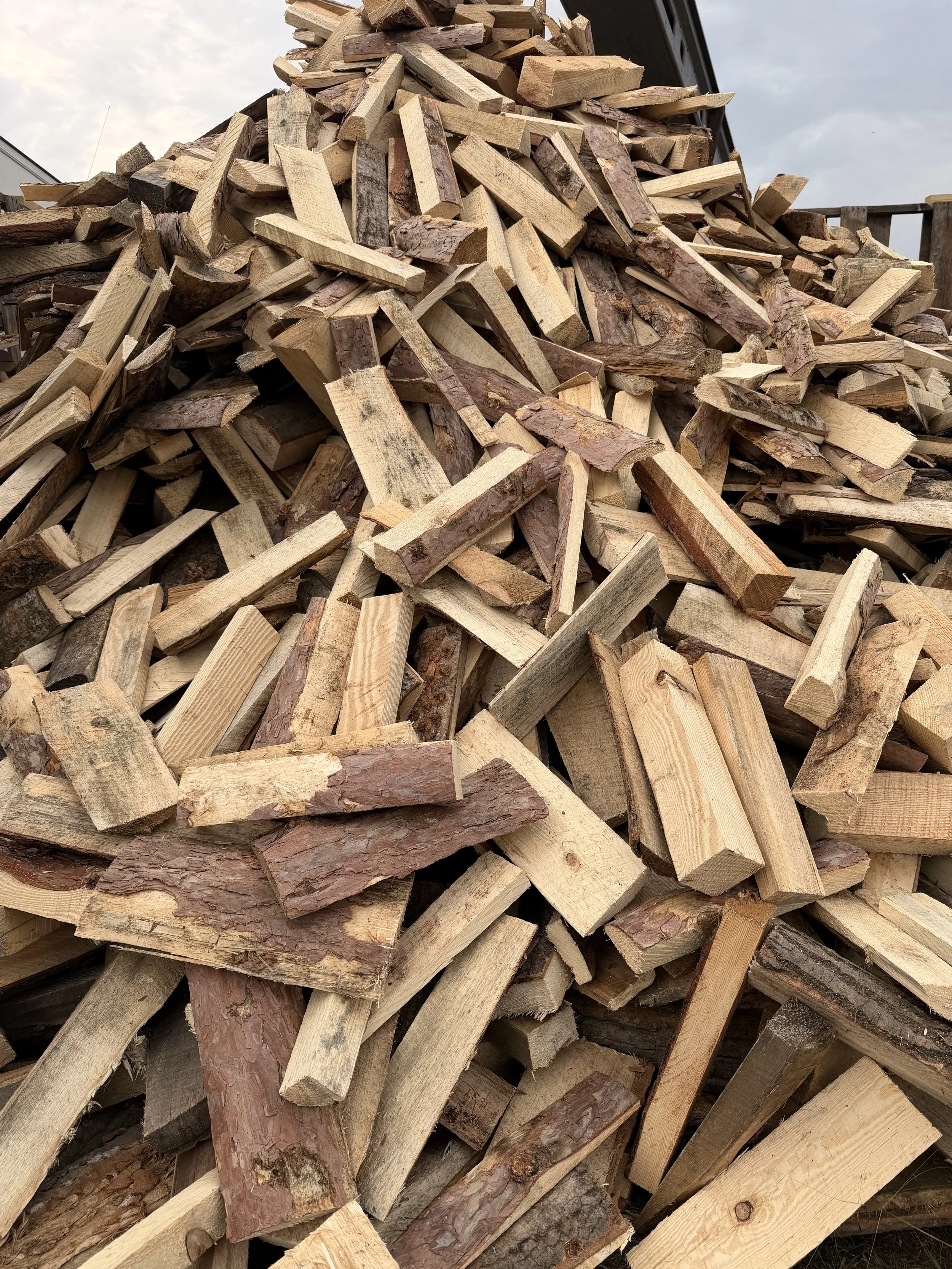 A large pile of chopped firewood stacked outdoors, with a partly cloudy sky in the background.
