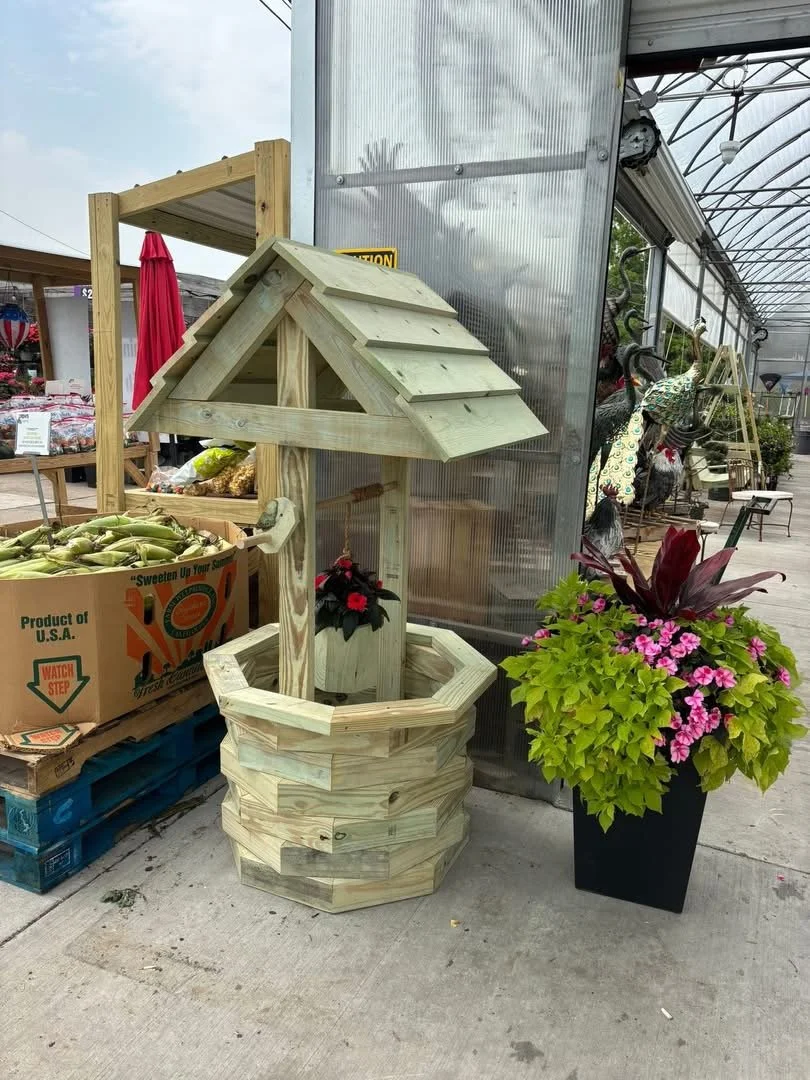 A wooden wishing well with a pitched roof, situated next to a large potted plant with pink flowers, outside a greenhouse or garden center.