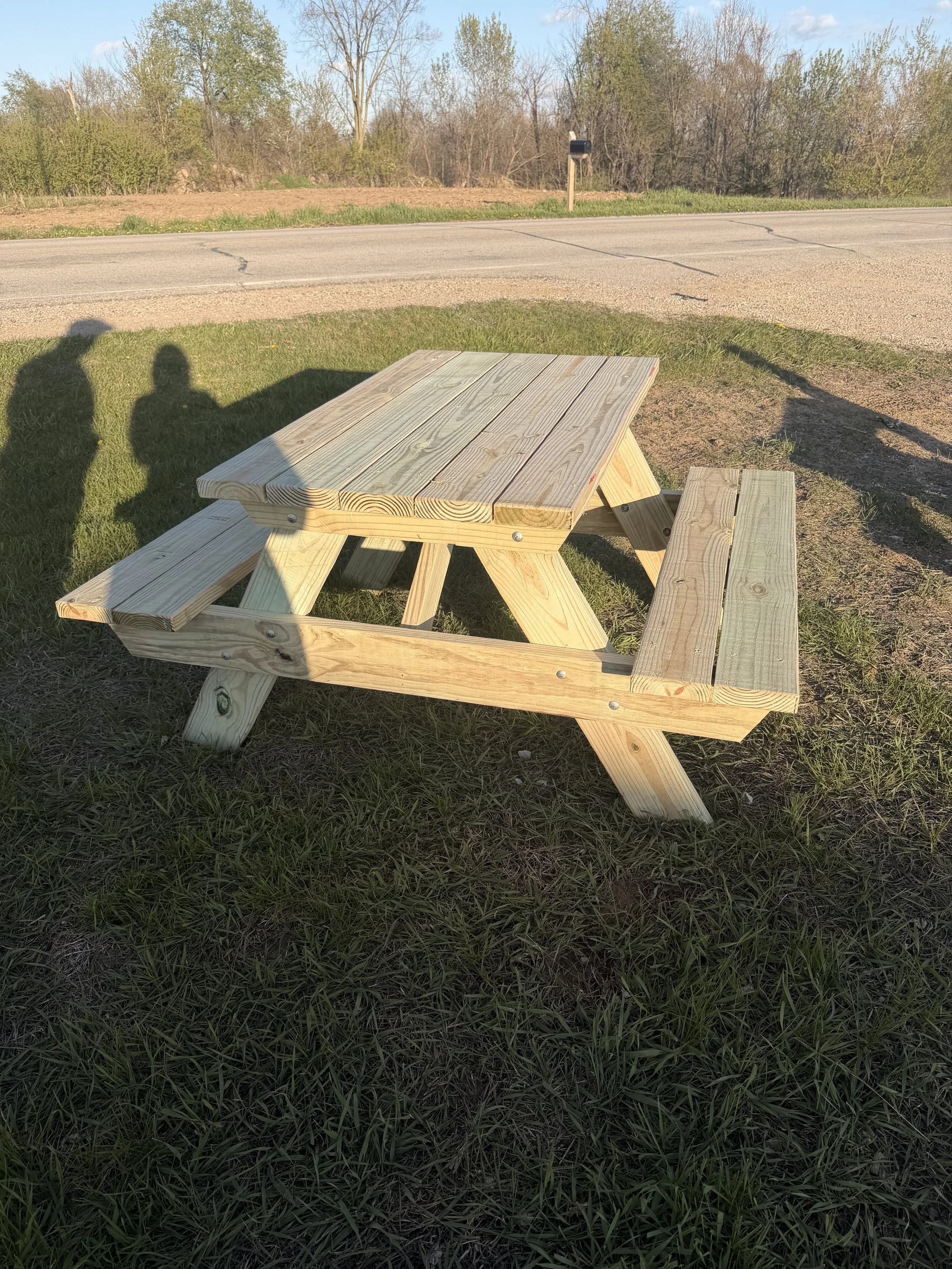 A wooden picnic table with attached benches positioned on a grassy area beside a paved road, during daylight with clear weather and scattered clouds, with trees in the background.