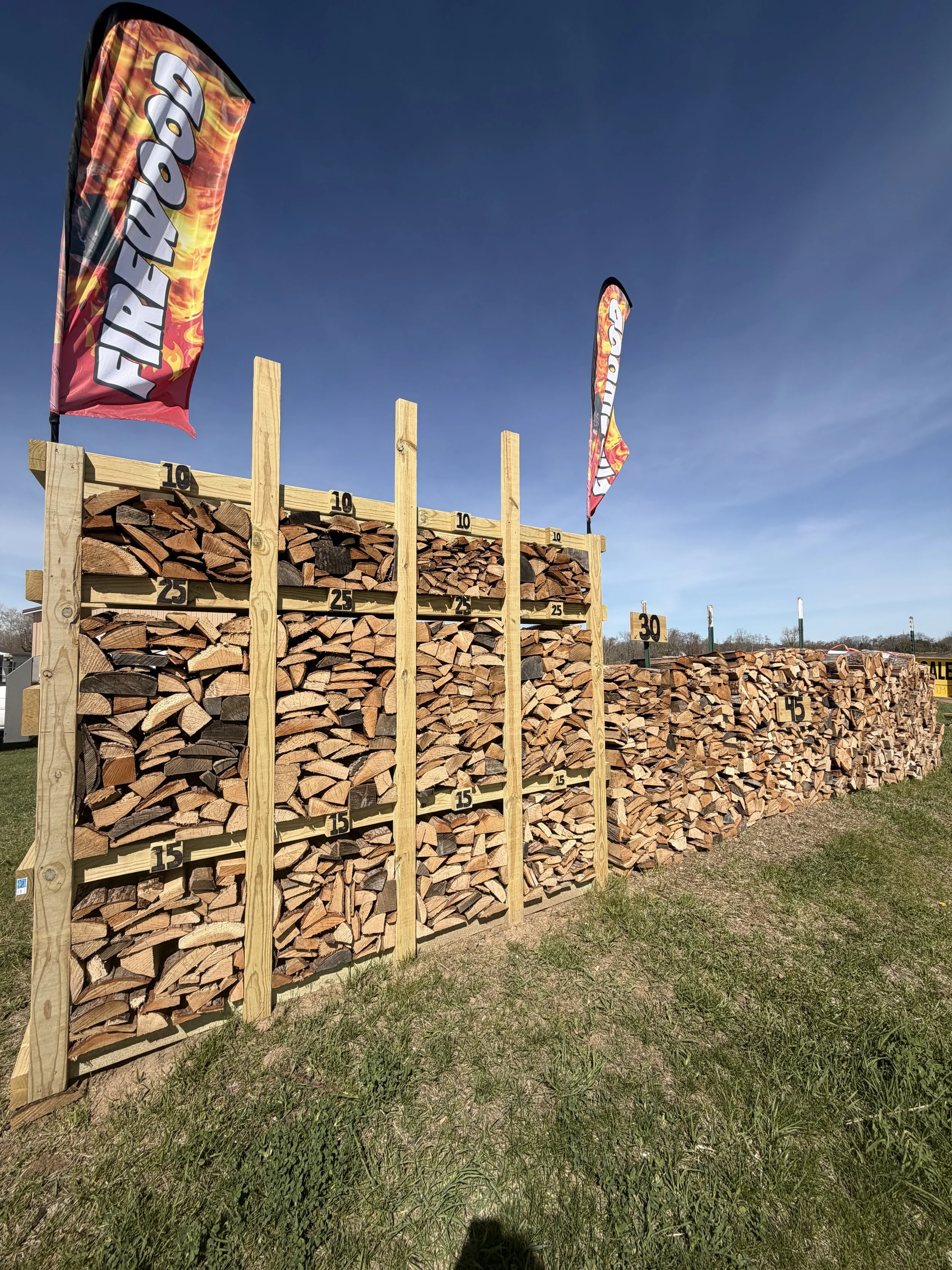 Stacks of firewood arranged on wooden racks with measuring labels, under a clear blue sky with two flags that read 'Firewood'.