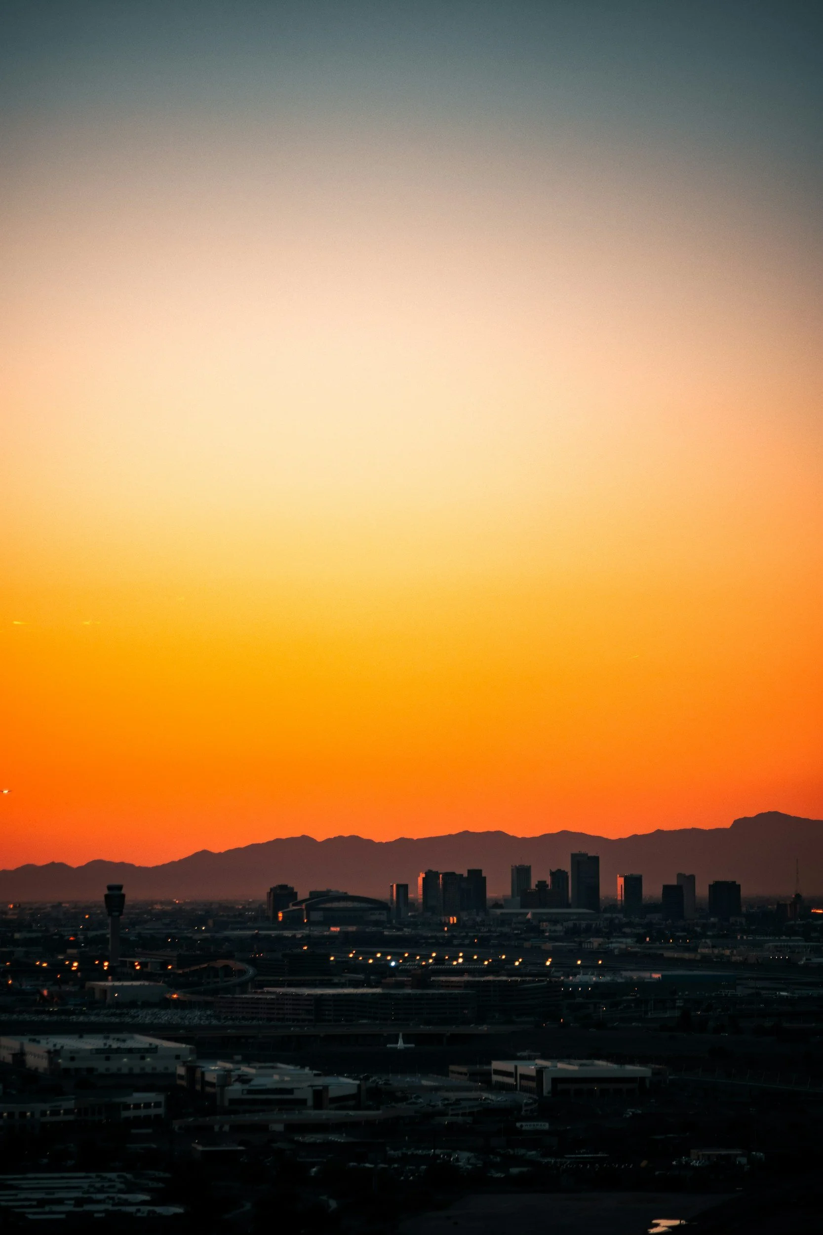 Sunset over a city skyline with mountains in the background, dark silhouettes of buildings in the foreground.