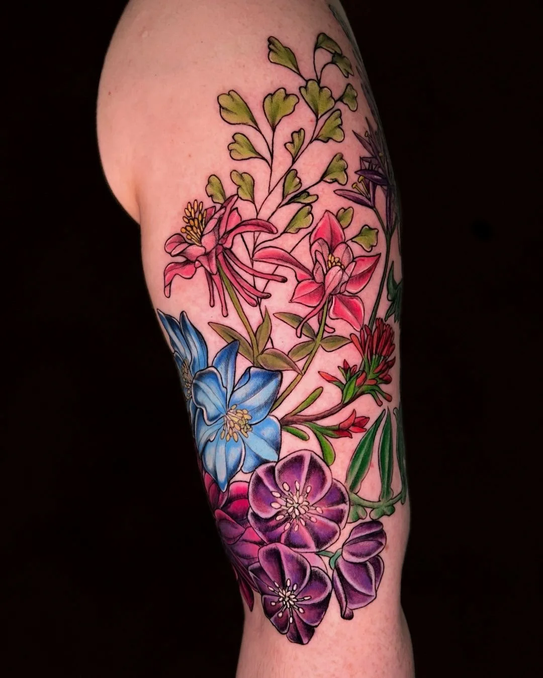 Colorful floral tattoo featuring pink, blue, purple, and red flowers with green leaves on a person's arm.