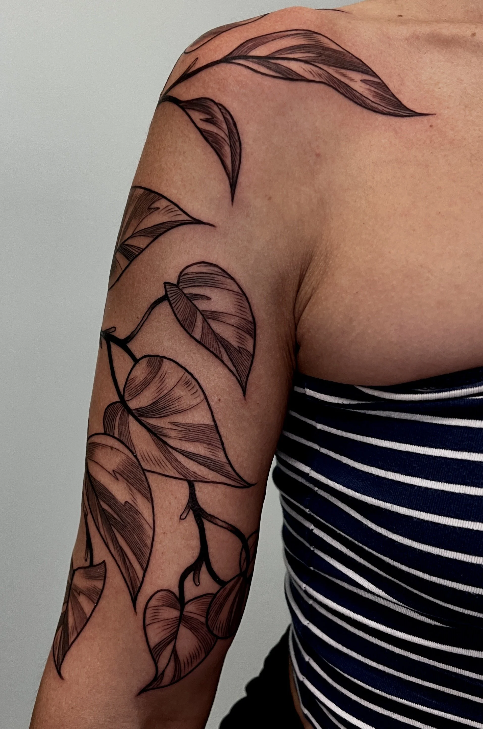 A person's arm with a black line art tattoo of leaves and stems, wearing a striped shirt.