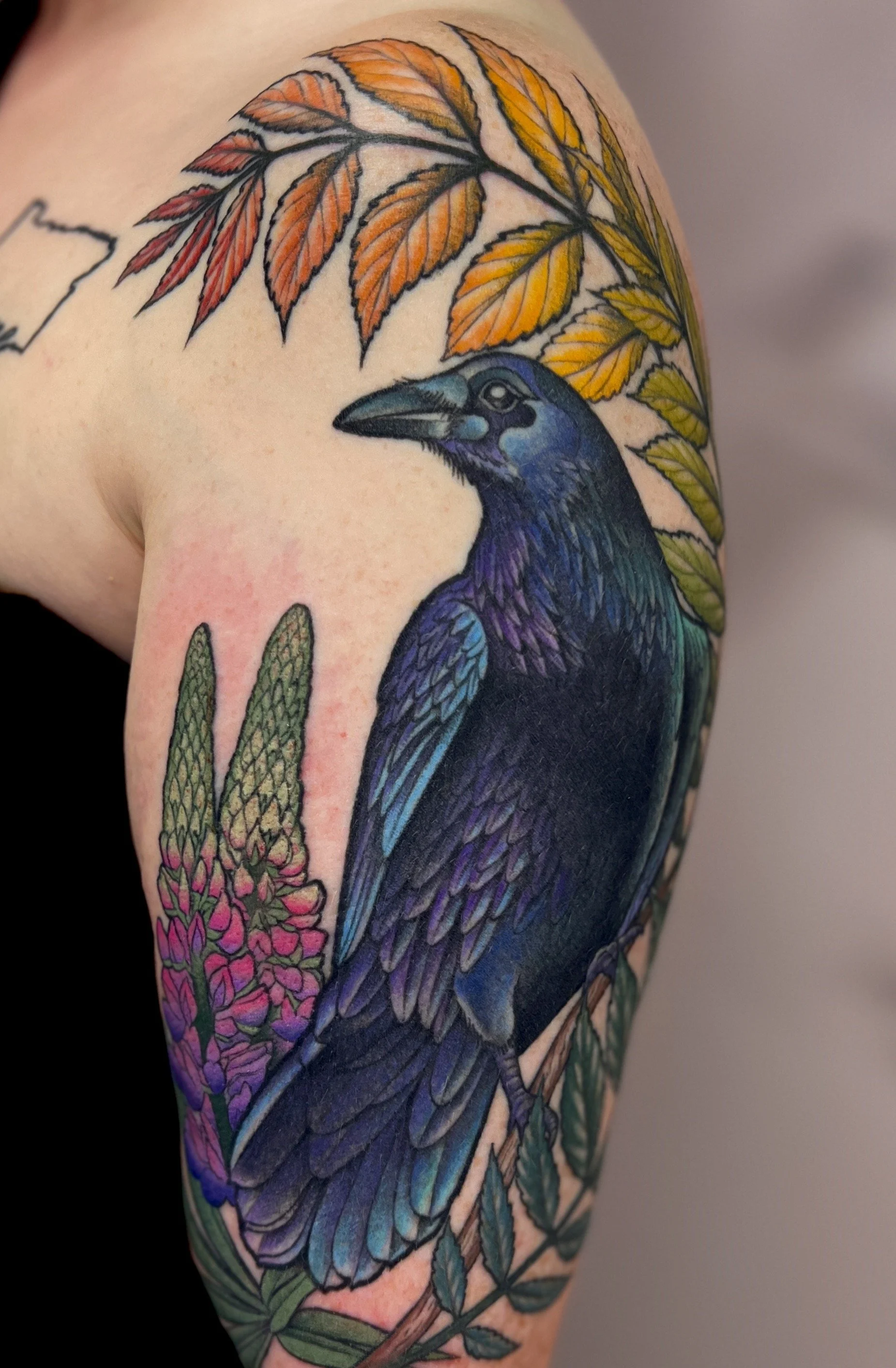 Colorful tattoo of a raven with orange and yellow leaves and pink flowers on the shoulder.