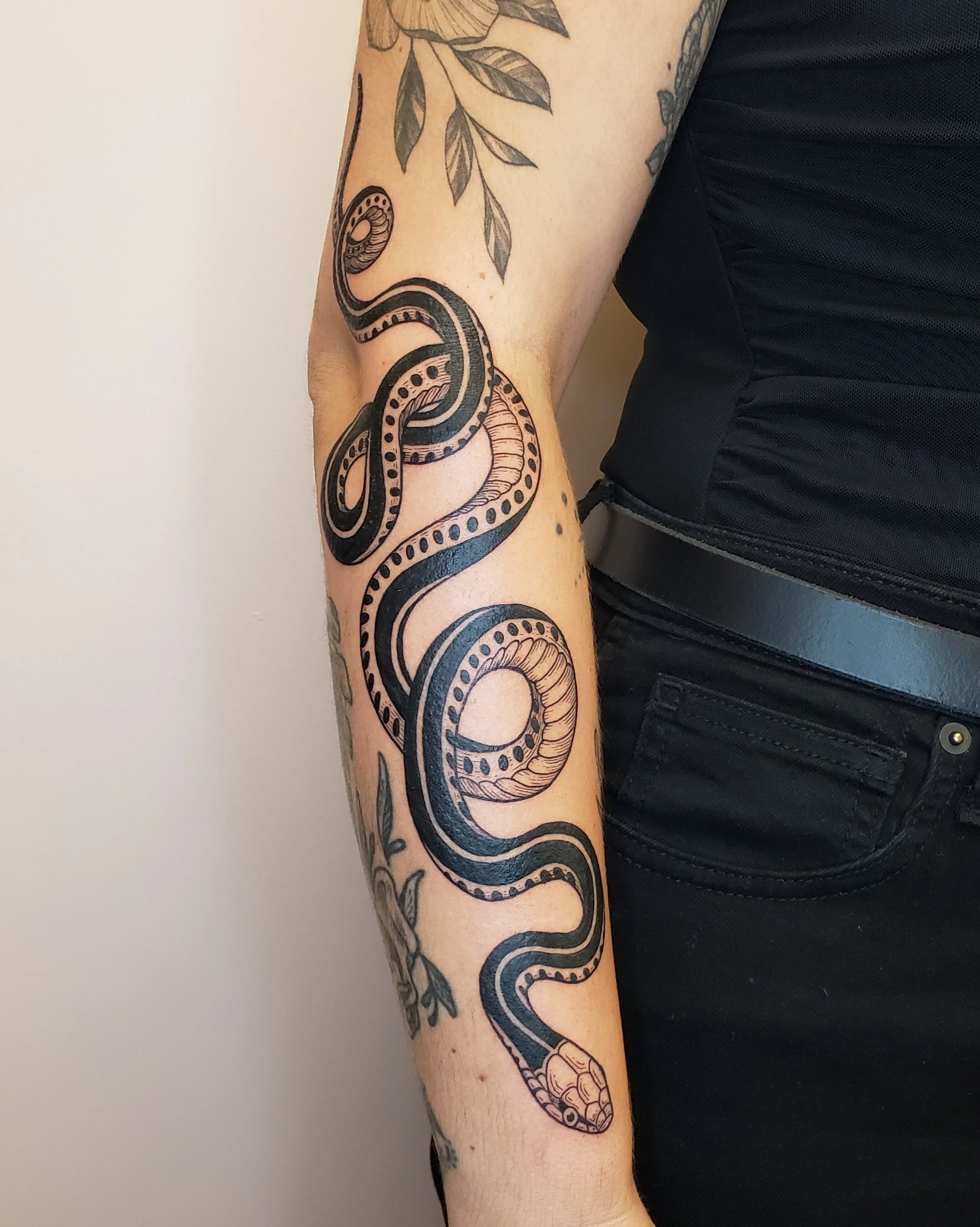 A tattoo of a snake with detailed scales and patterns, coiled around the person's arm, with leaves above it. The person is wearing a black short-sleeve shirt.