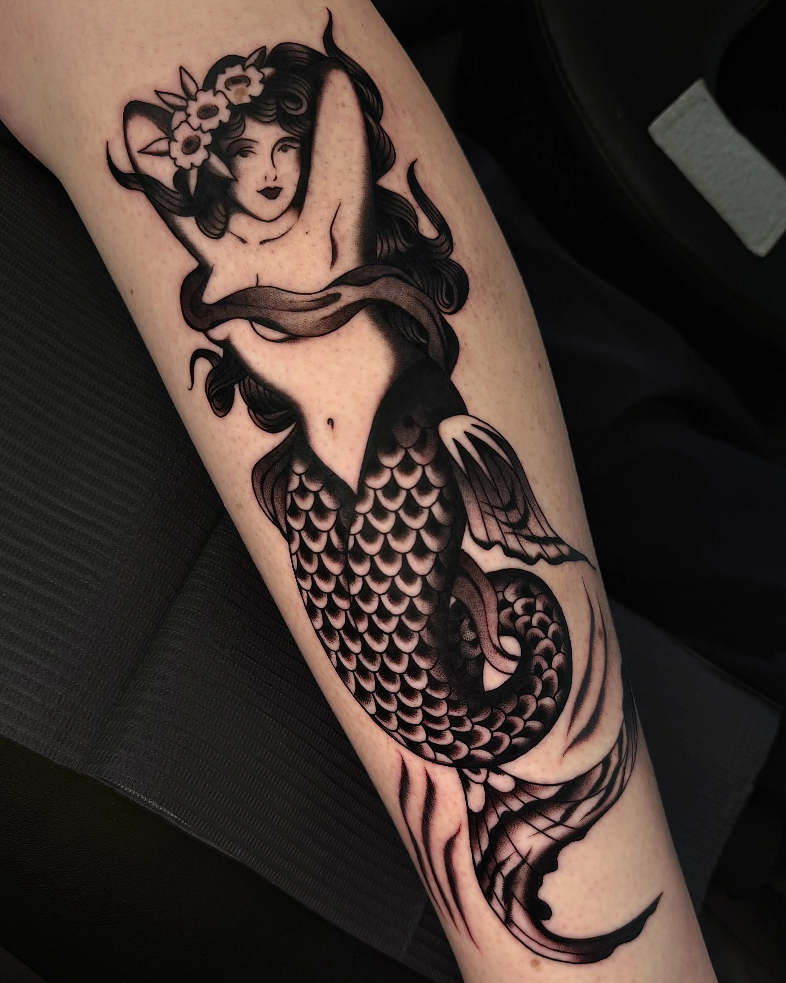 Black and gray tattoo of a mermaid with long flowing hair, a flower crown, and a fish tail, on a person's arm.